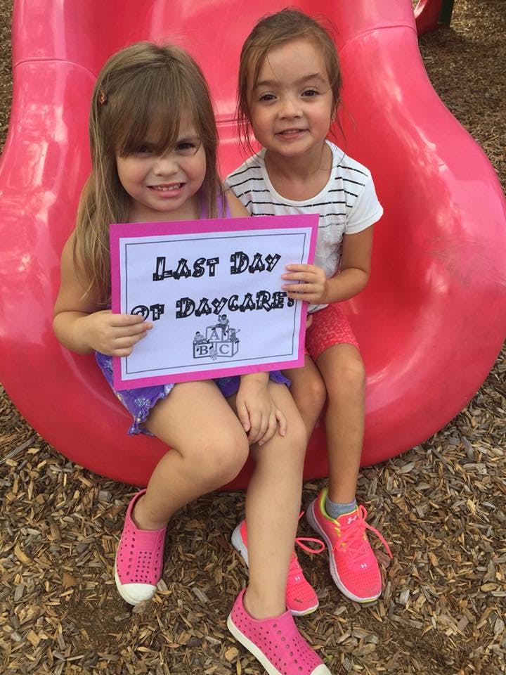 Two children sitting on a red slide holding a "Last Day of Daycare" sign