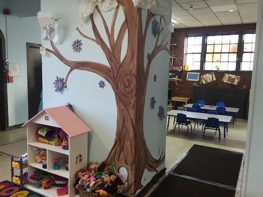 Tree mural on a wall next to a dollhouse and a classroom with tables and chairs