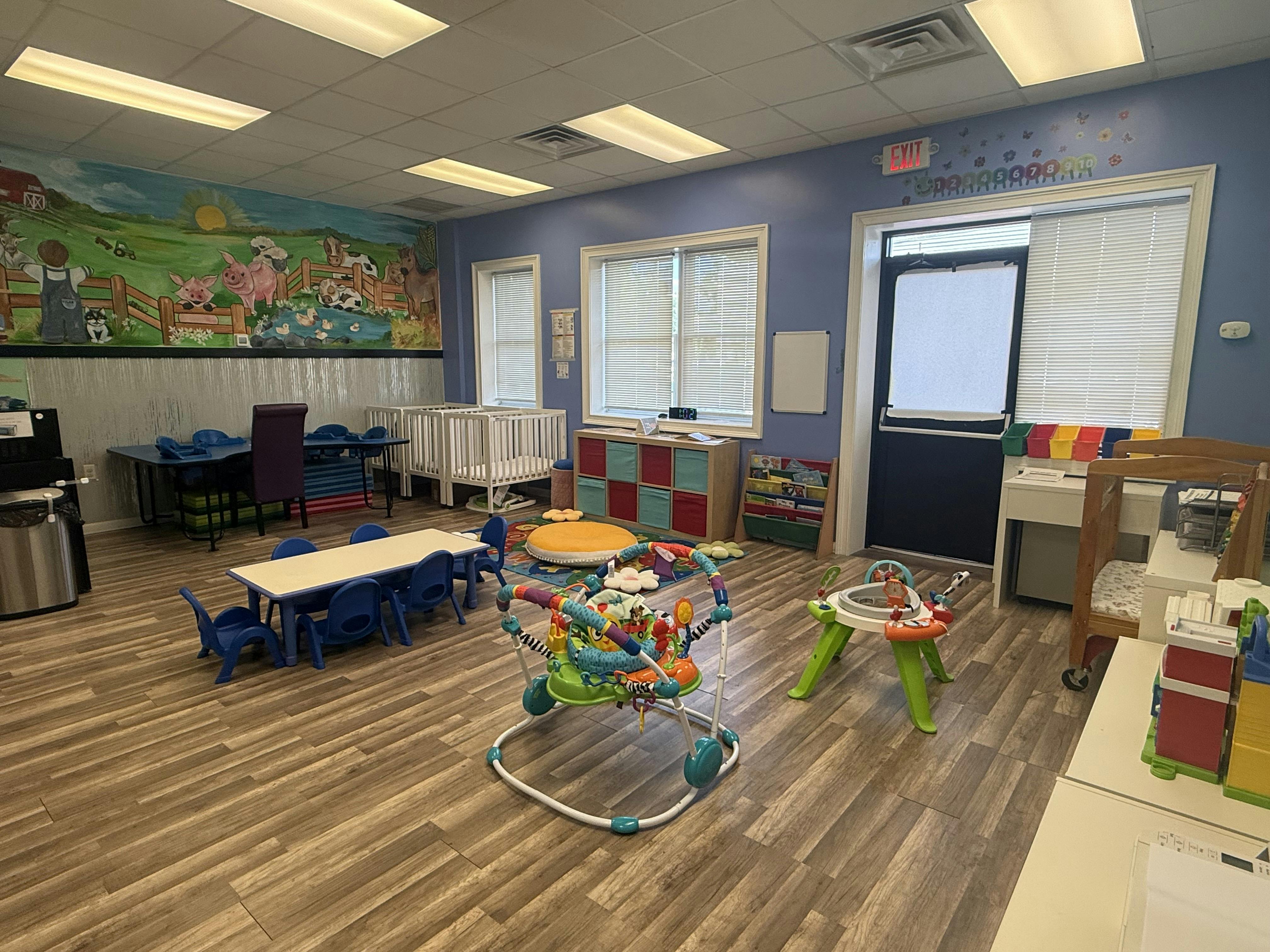 Childcare room with baby activity centers, small tables, chairs, and cribs