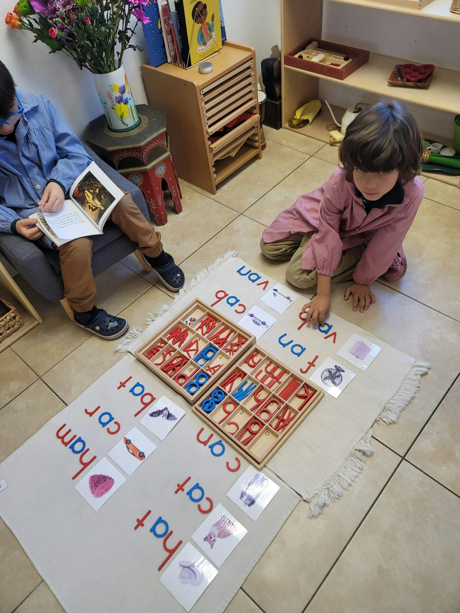 Child on floor with letter tiles and word cards, person reading in background