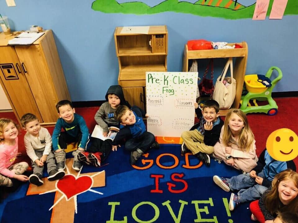 Children sitting on a rug with "God is Love" text, a whiteboard behind them