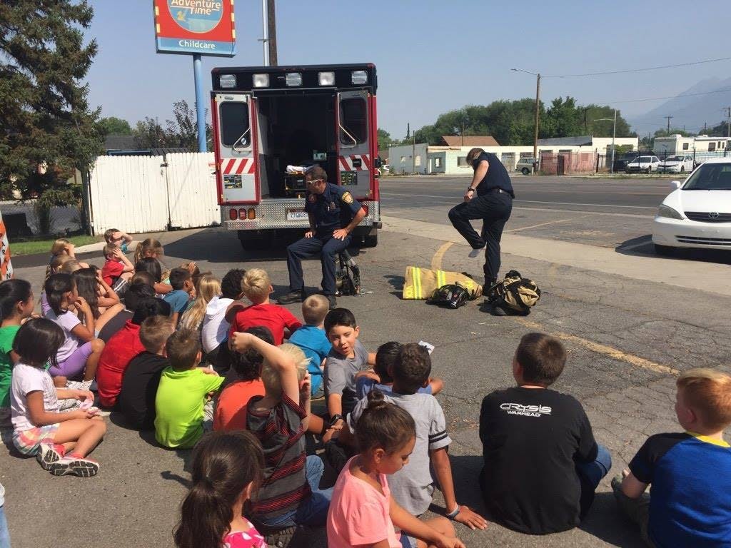 Children sitting on the ground watching two people in uniform near an ambulance