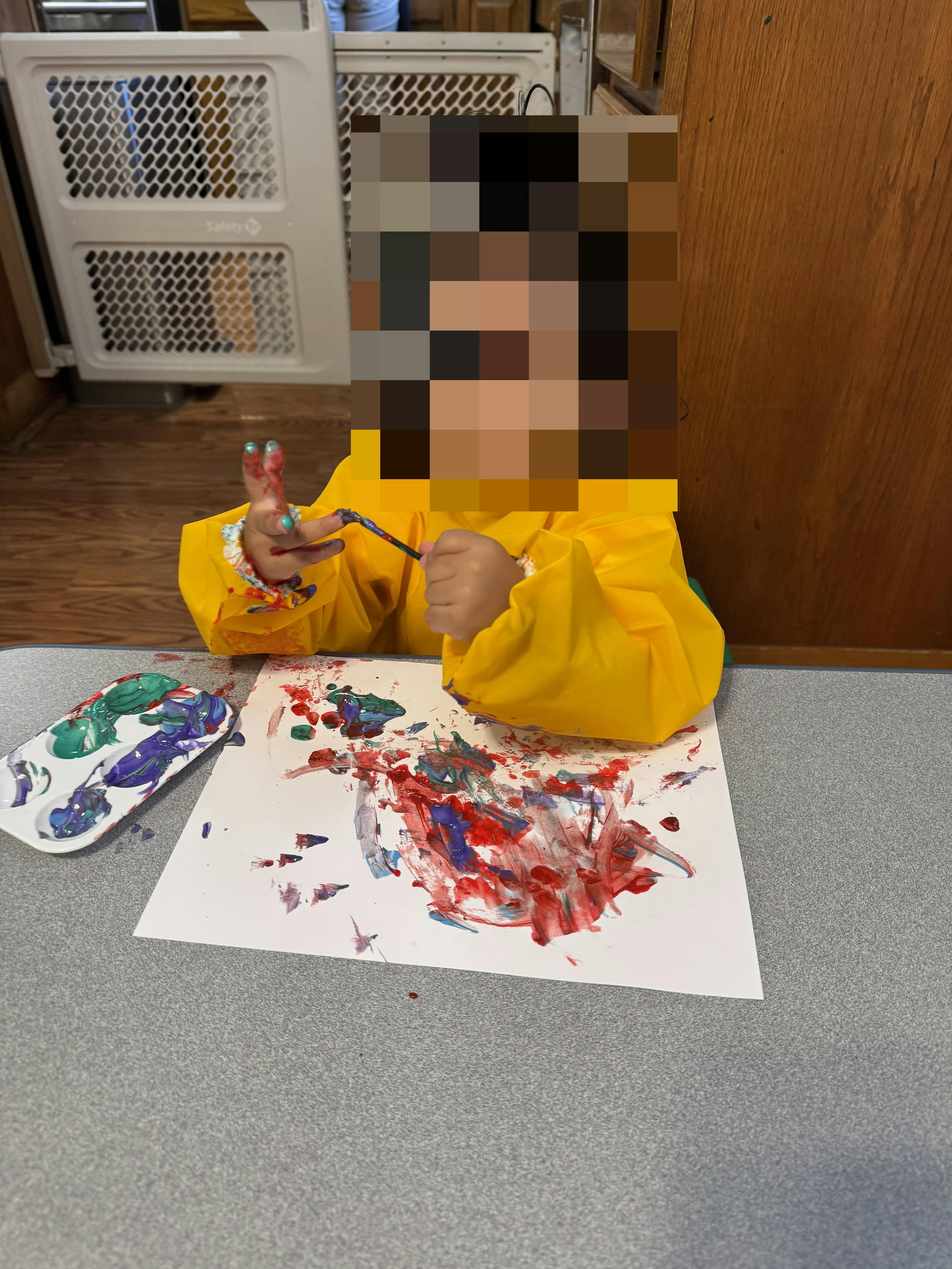 Child wearing a yellow smock, painting with fingers and a brush at a table