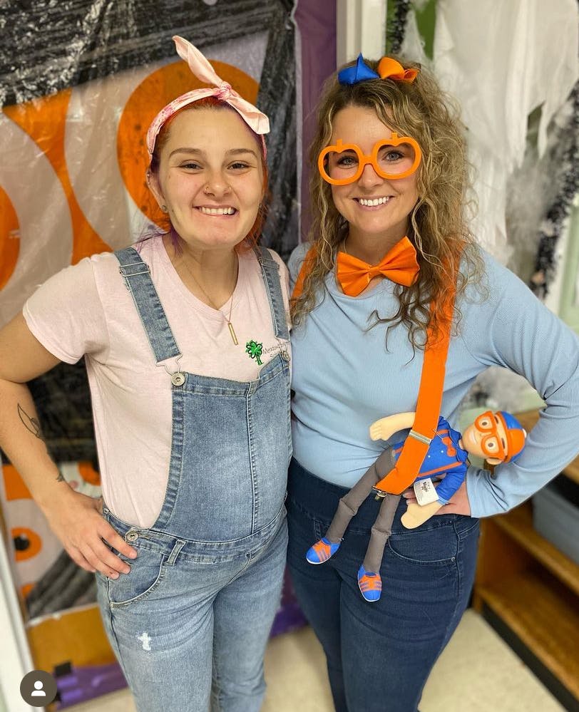 Two people smiling, one in overalls, one in orange glasses and suspenders