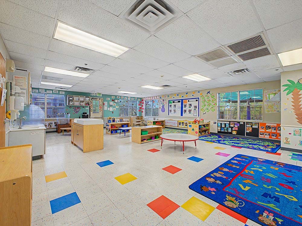 A colorful empty classroom with small tables, chairs, and play mats on the floor