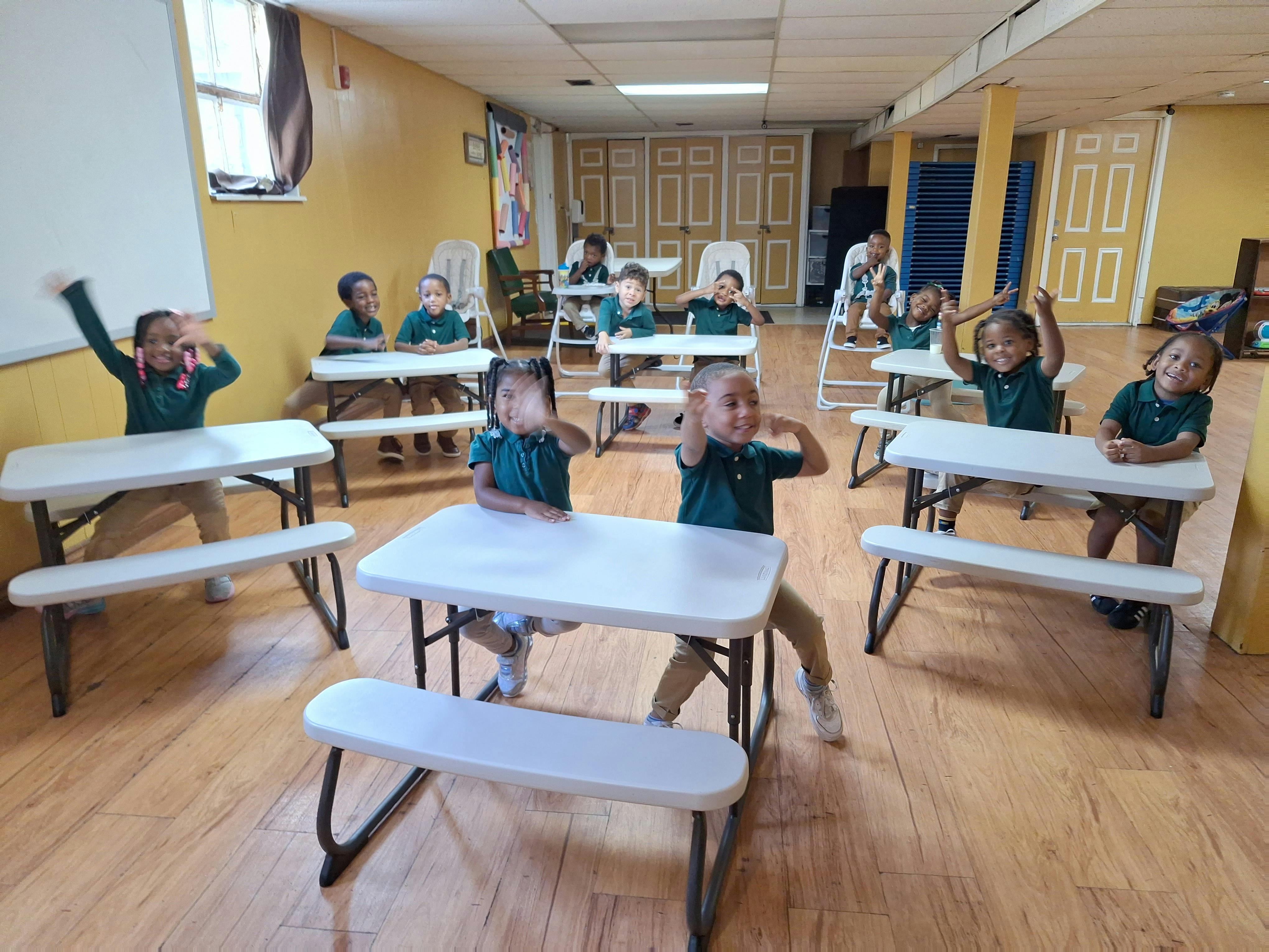 Children sitting at tables in a classroom, some raising their hands