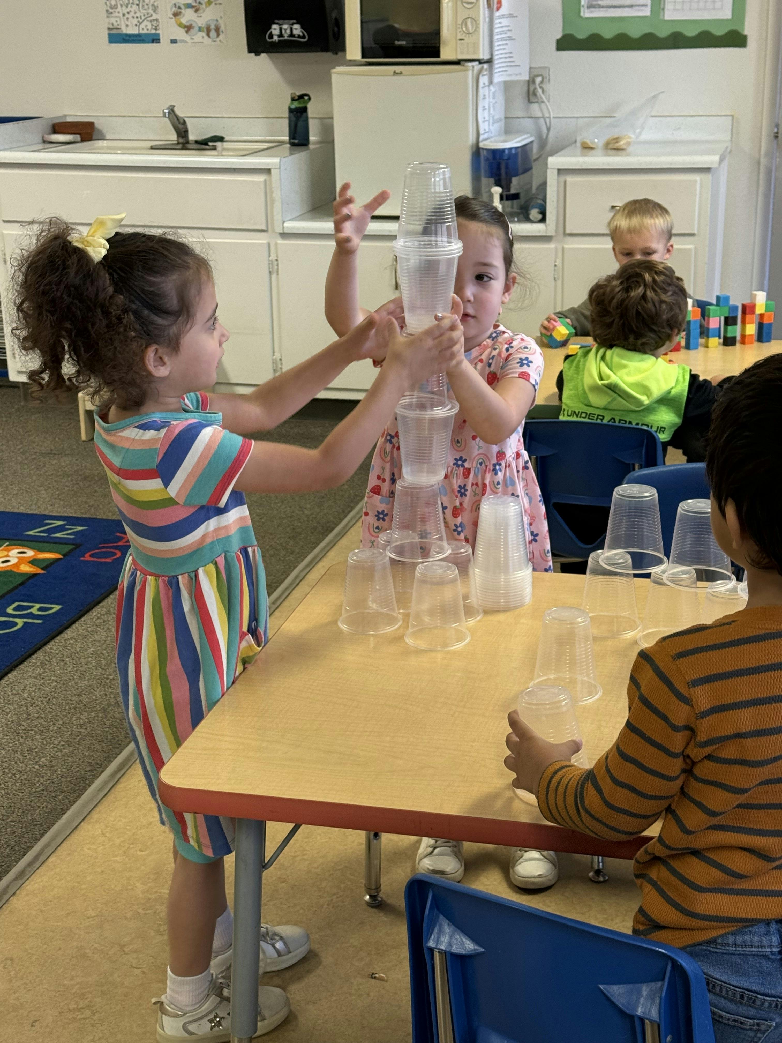 Two children stacking plastic cups in a classroom, with other children in the background