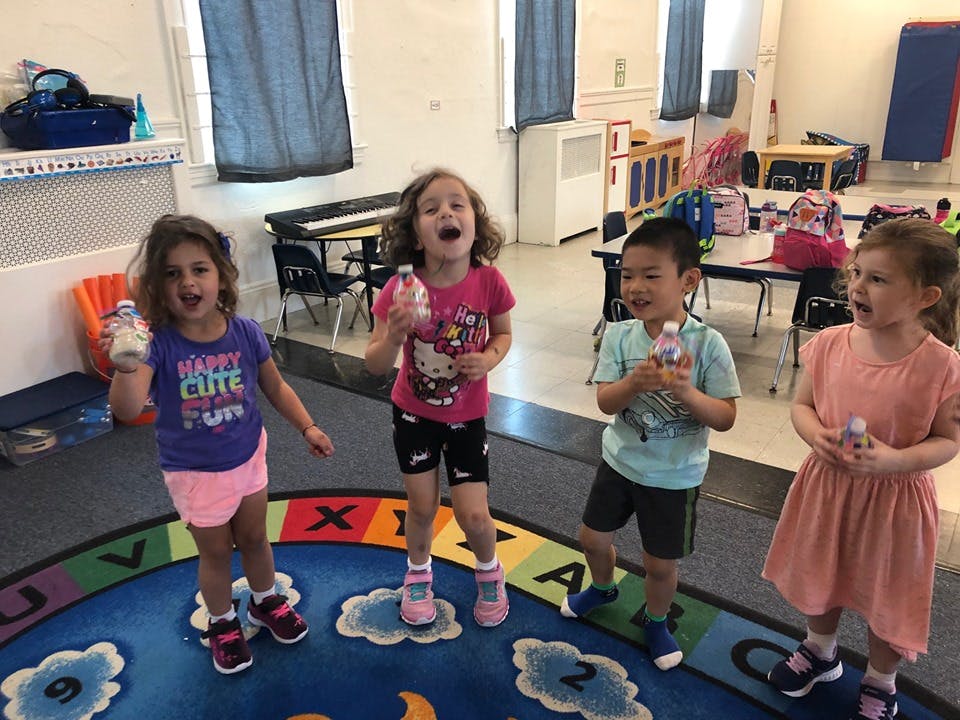 Four children holding bottles and standing on a rug in a classroom