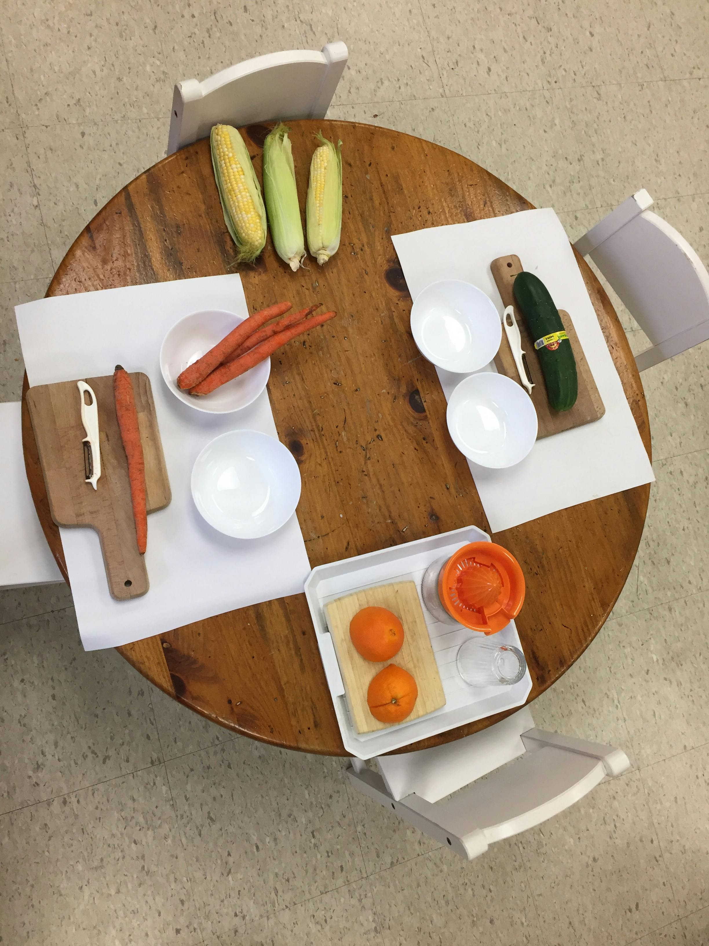 Table with corn, carrots, cucumber, oranges, bowls, and cutting boards