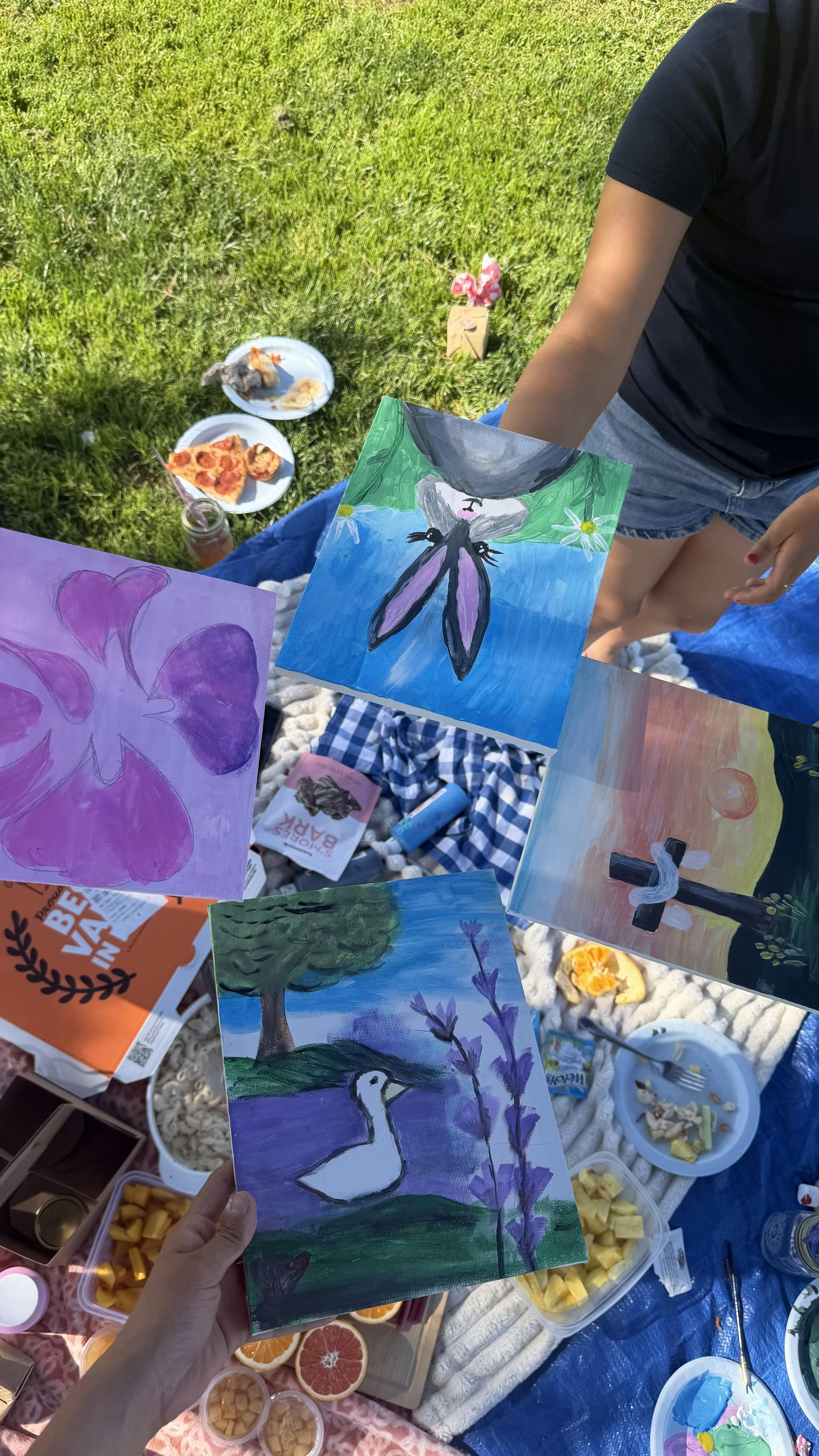 Multiple people holding up their paintings at a picnic in the grass