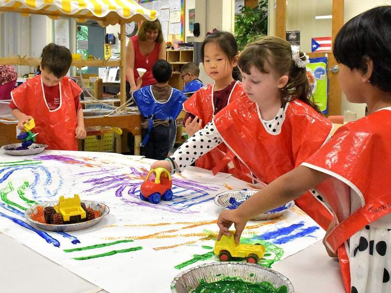 Children in smocks painting with toy cars on a large sheet of paper
