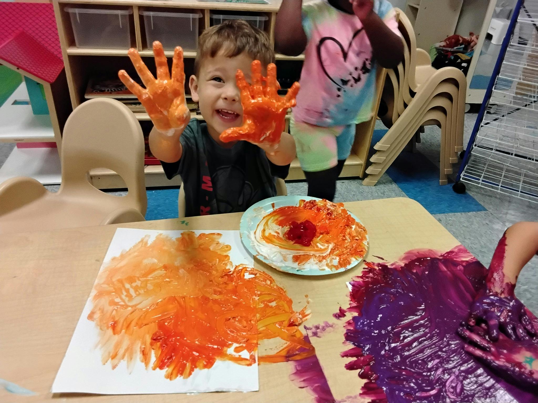 Child with orange paint on hands, another child's hands in purple paint