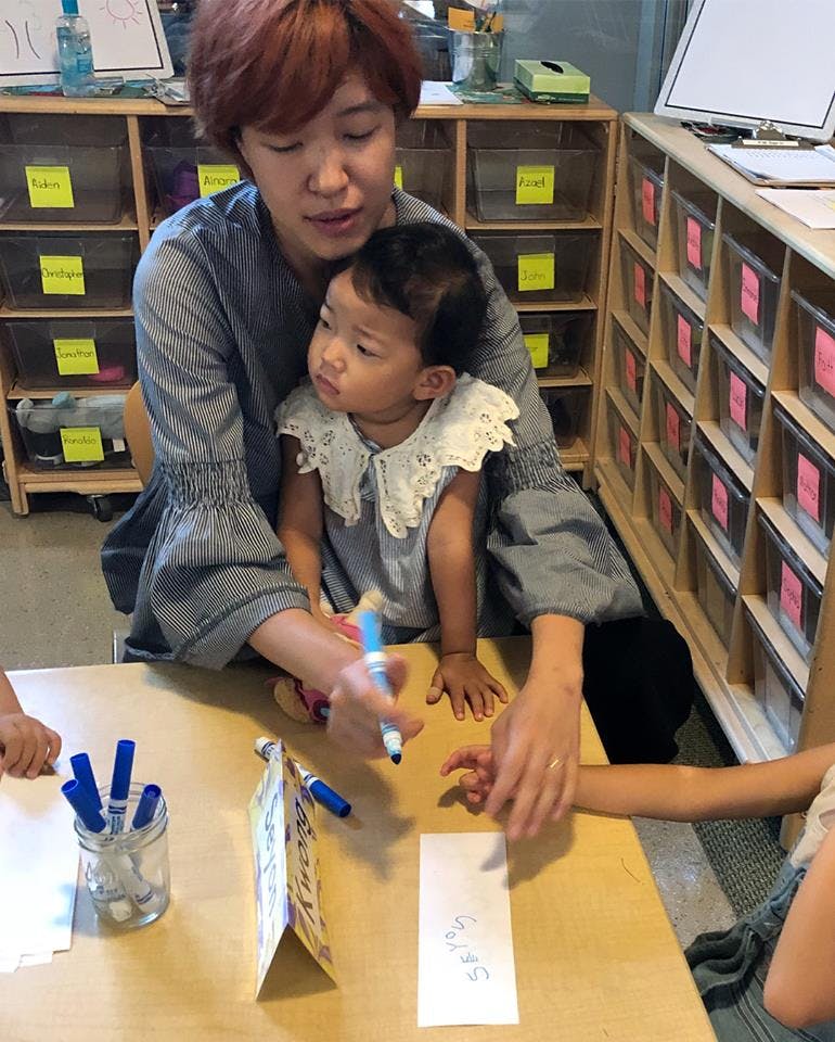 Person holding a child while another person writes on paper with a marker