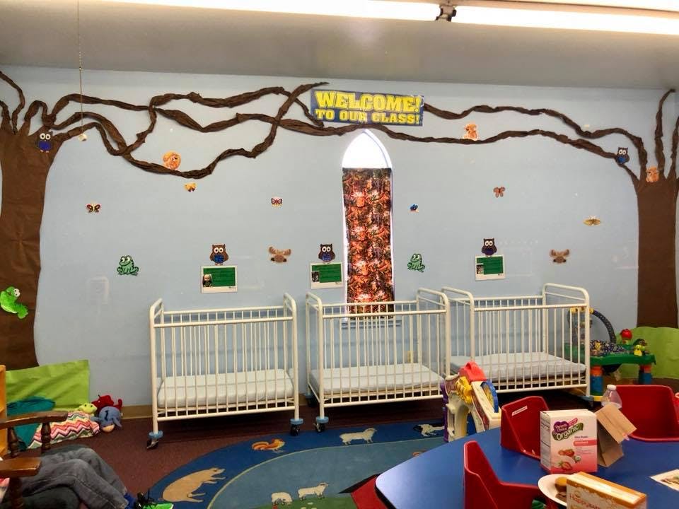 Three white cribs in a room with a tree mural and "WELCOME TO OUR CLASS!" sign