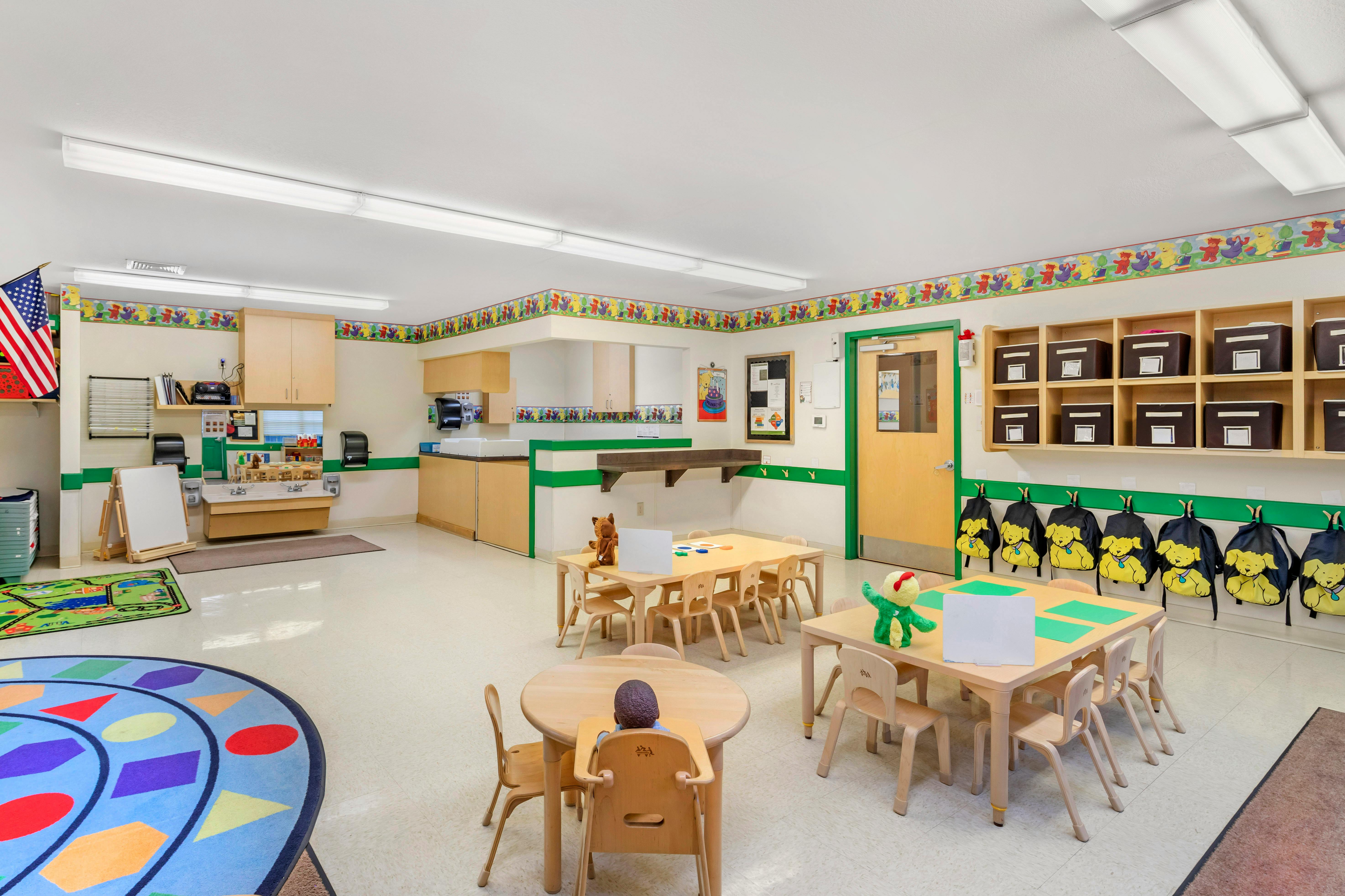 A classroom with small tables and chairs, a sink area, and cubbies with backpacks