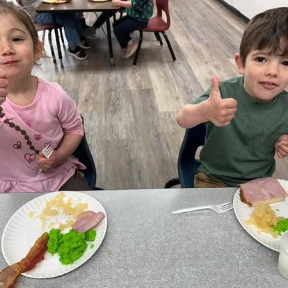 Two children sitting at a table giving a thumbs up gesture while eating lunch