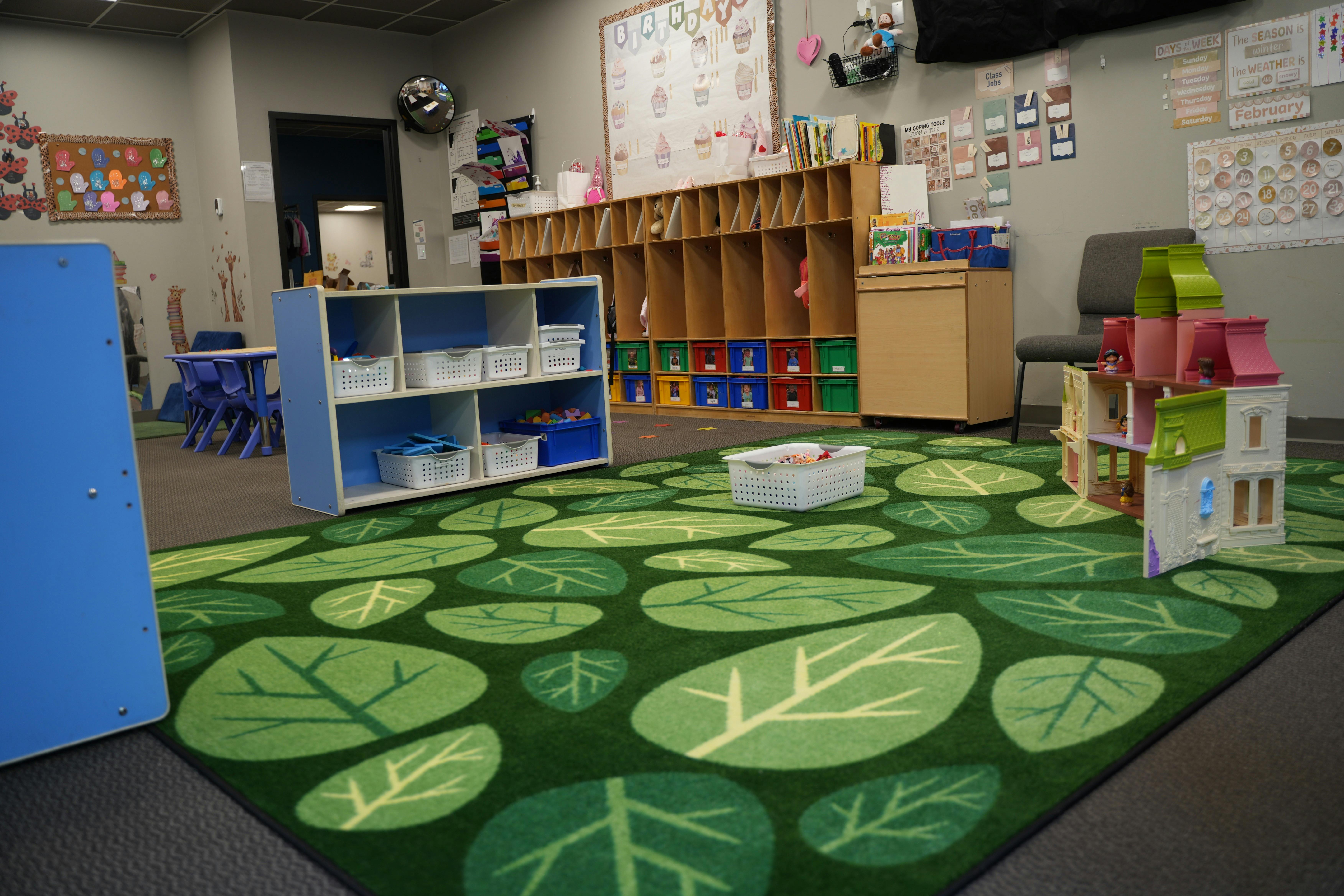 Classroom with green leaf rug, shelves, cubbies, and a dollhouse