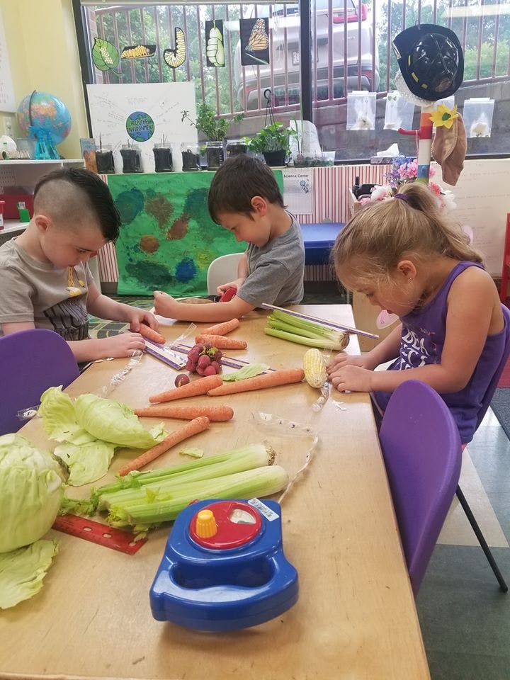 Three children peeling vegetables at a table in a classroom