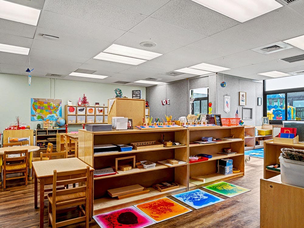 Classroom with small wooden tables and chairs, shelves, and educational materials