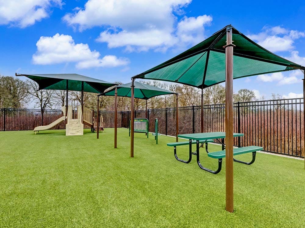 A playground with a slide, shades, and picnic tables on artificial turf