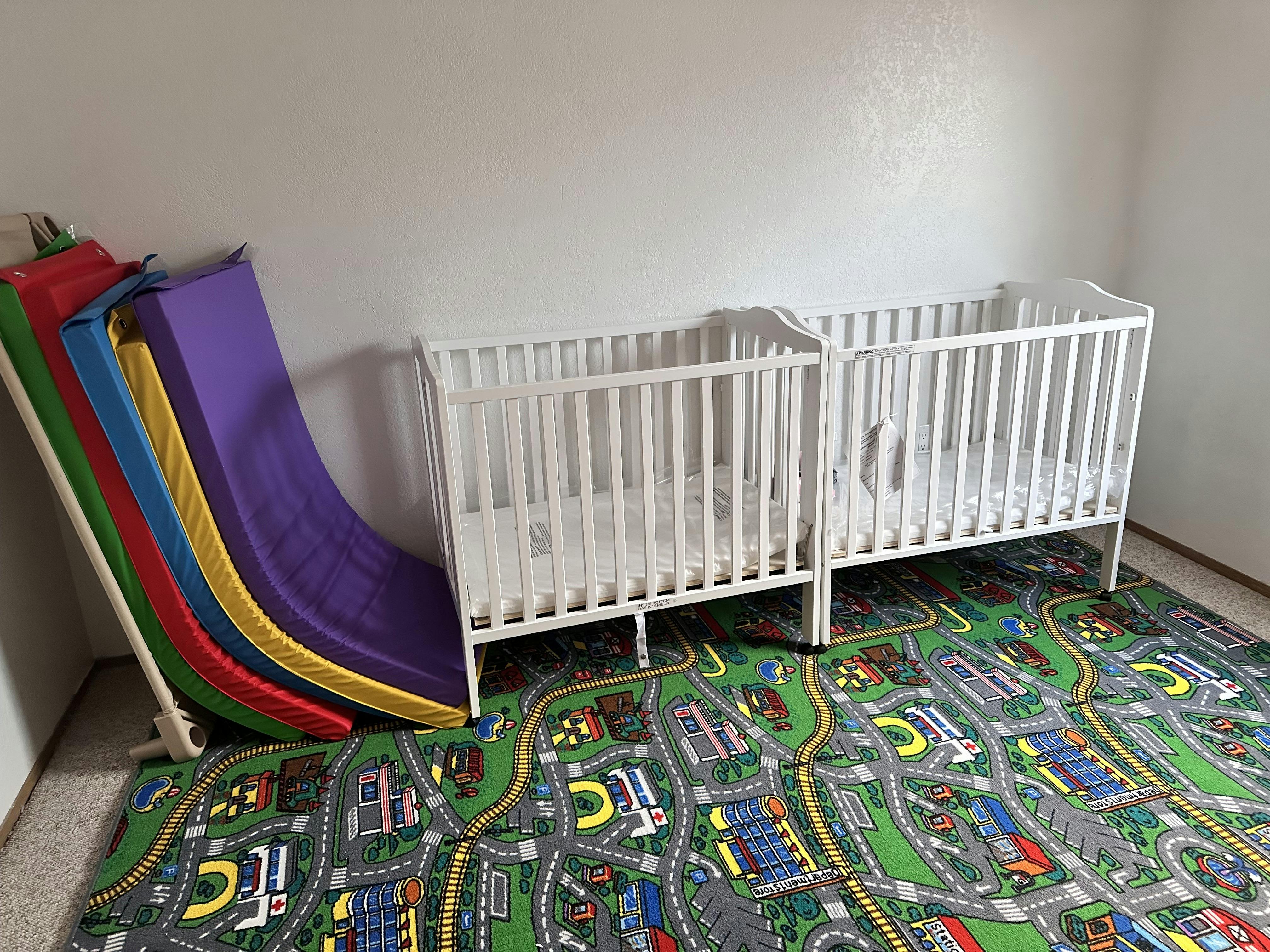 Two white cribs next to a colorful play slide on a road map rug