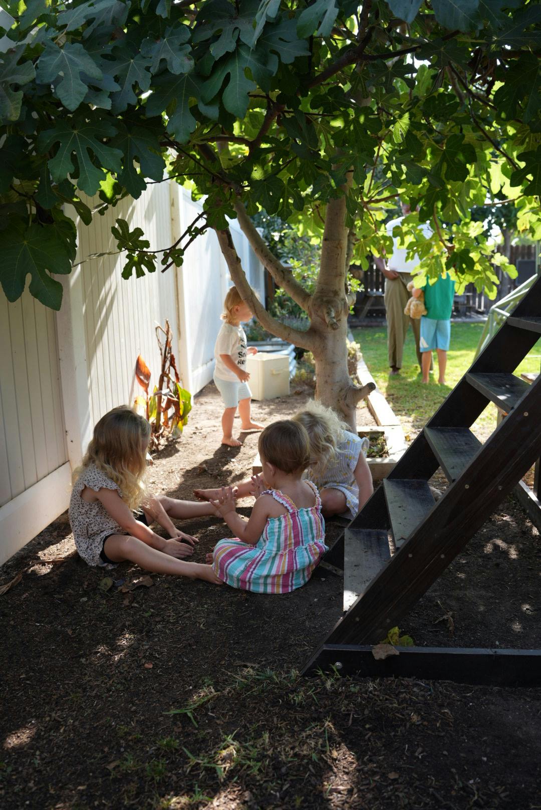 Three children playing on the ground near a house wall and tree