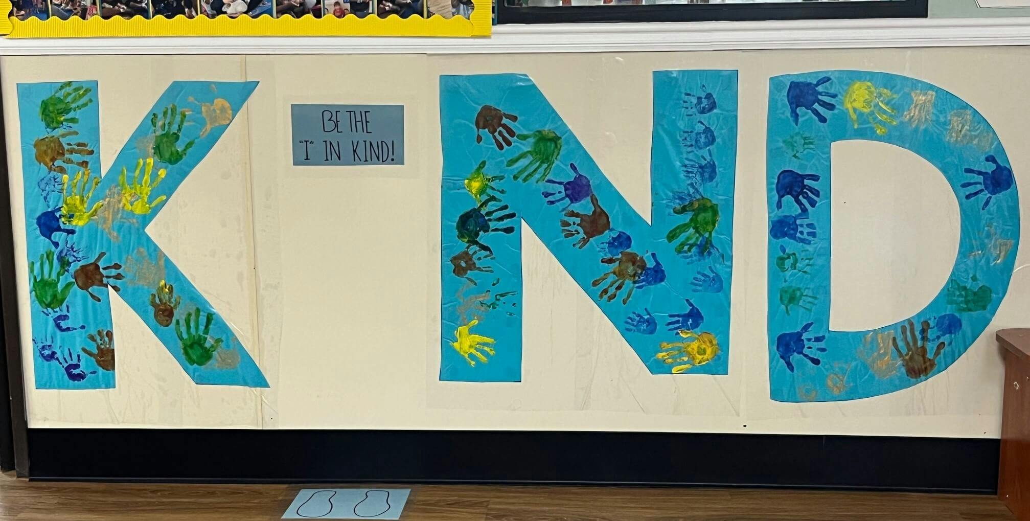 Classroom bulletin board with the word KIND made from blue paper and handprints