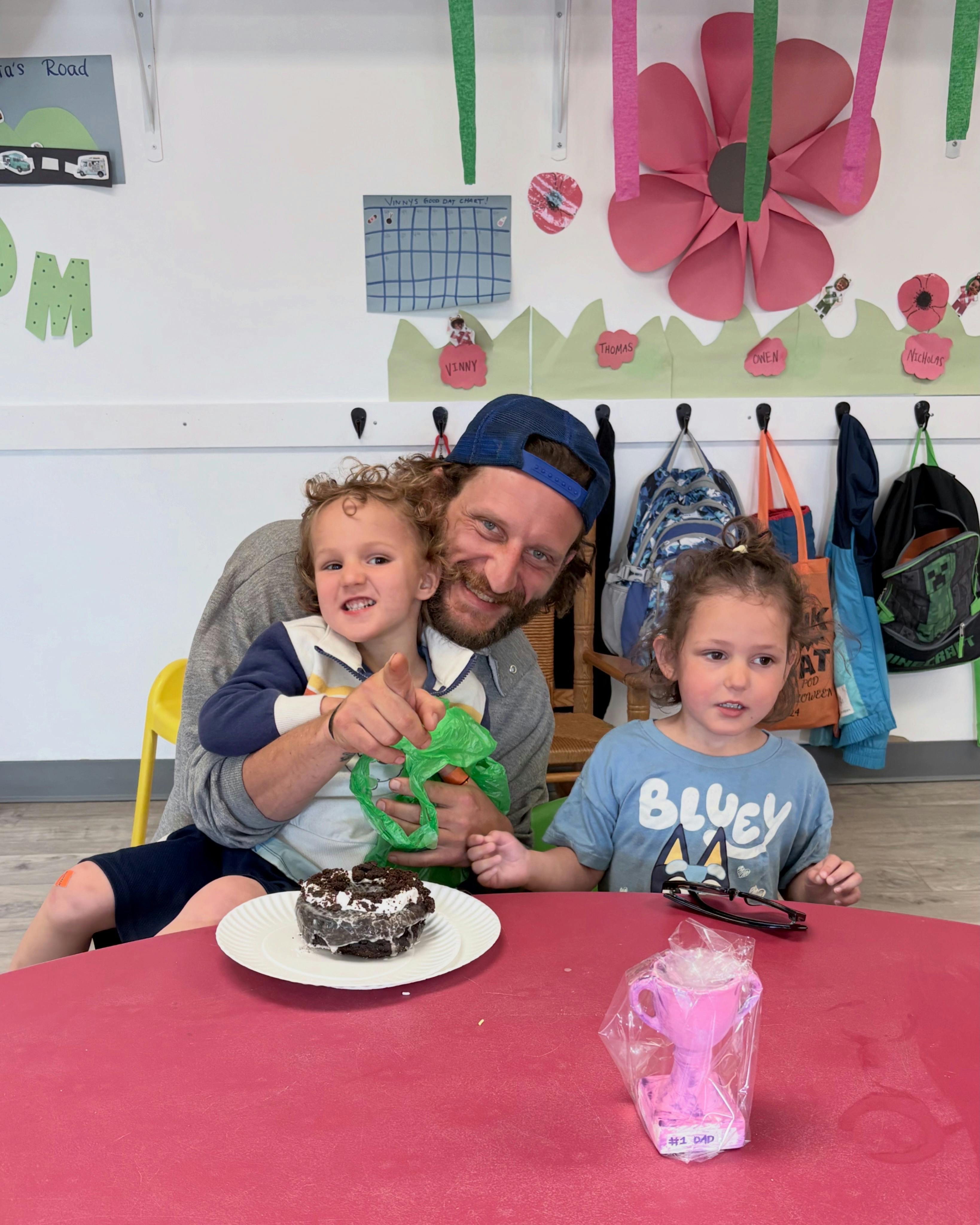 A person holding a child with another child sitting beside a desk with a donut
