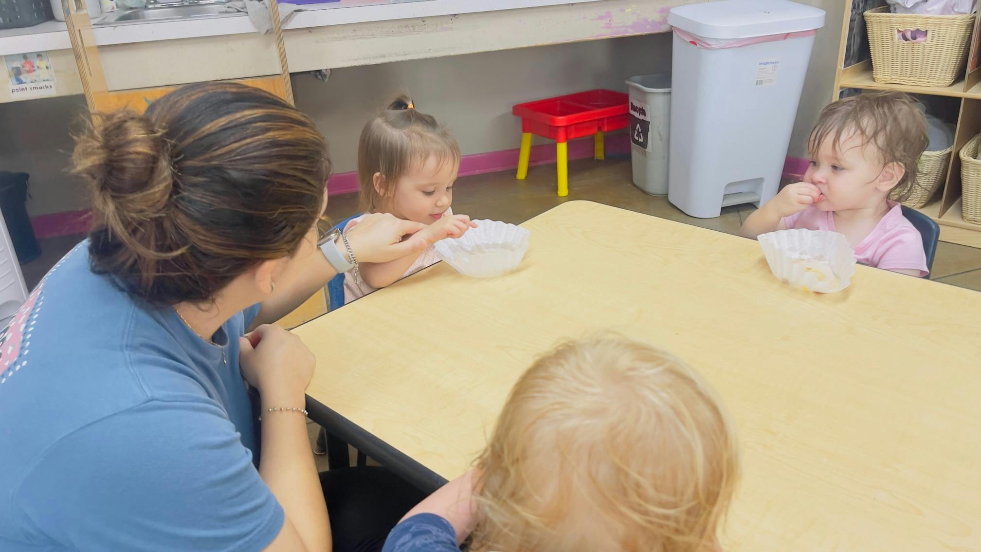 Person helping a child eat from a bowl at a table with other children