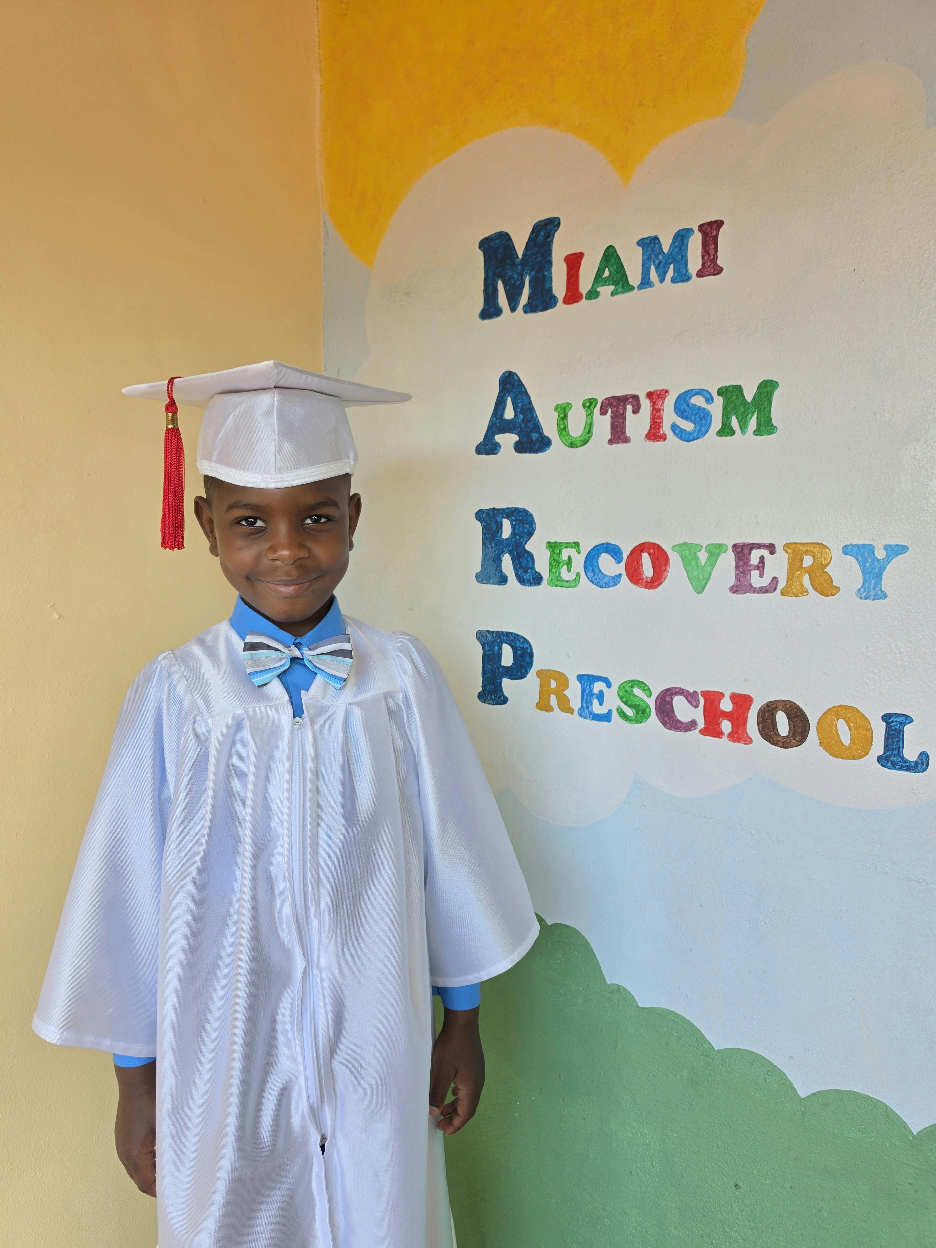 Child in a graduation cap and gown next to a sign for Miami Autism Recovery