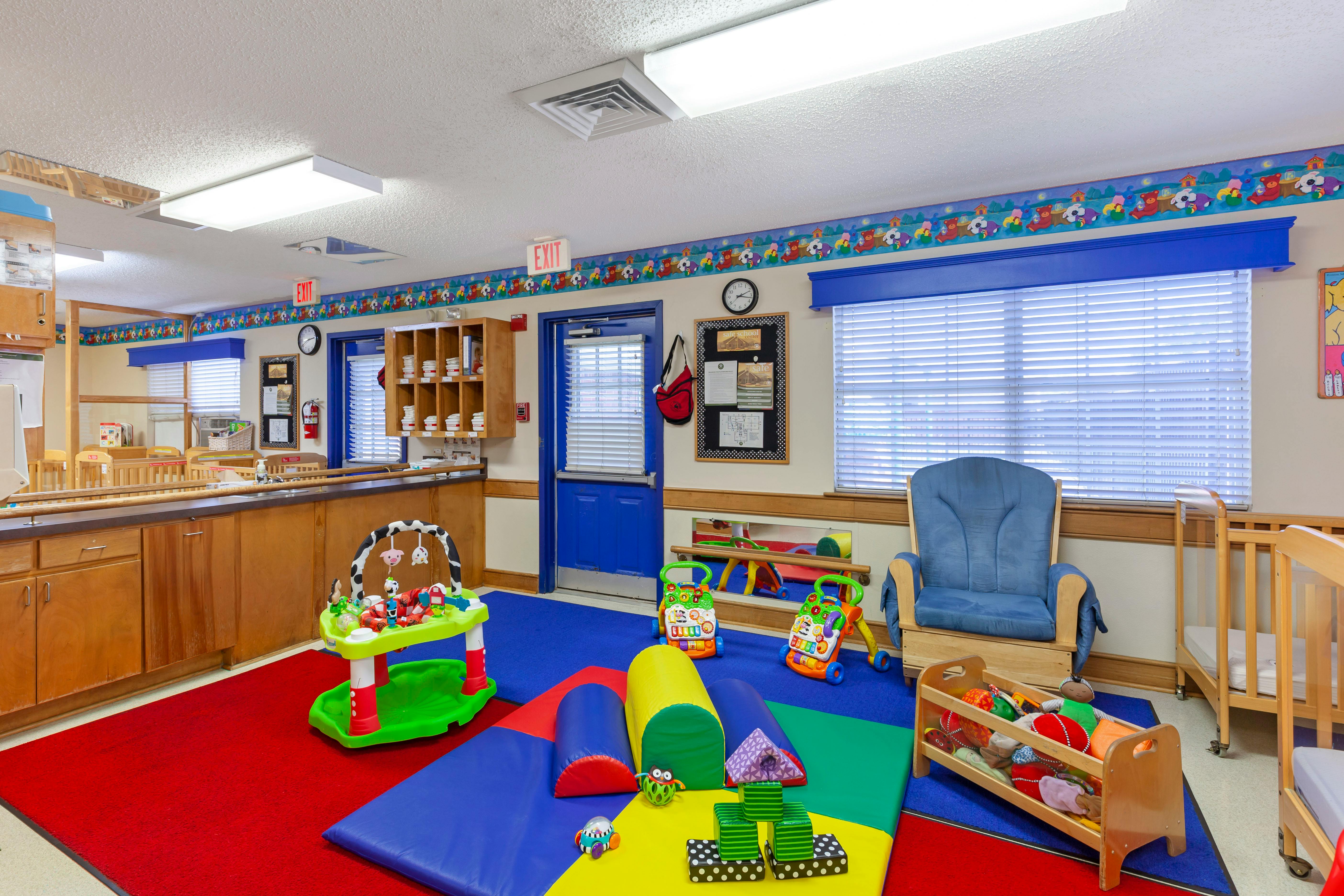 Children's classroom with colorful playmats, toys, blue door, and wooden cribs