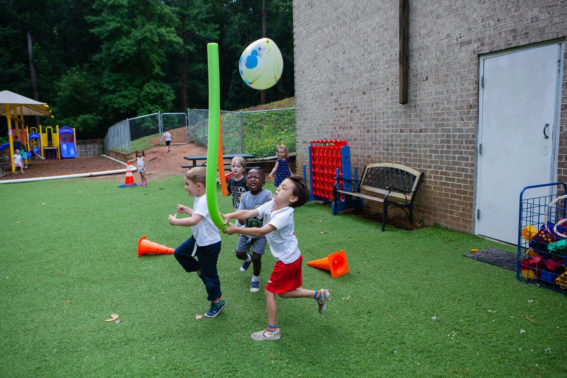 Children playing with a long green pool noodle and a large ball outdoors