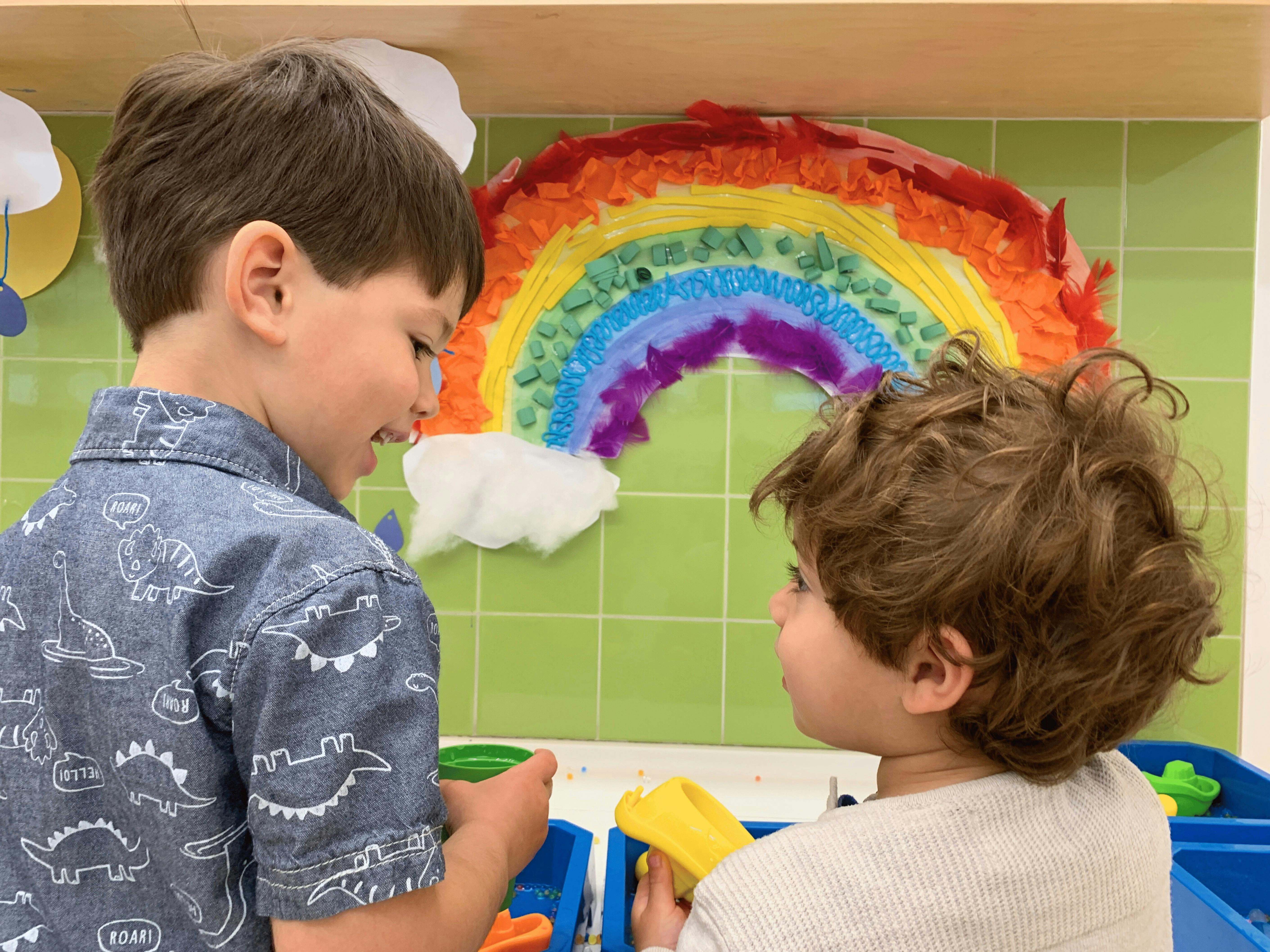 Two children looking at each other in front of a rainbow art project