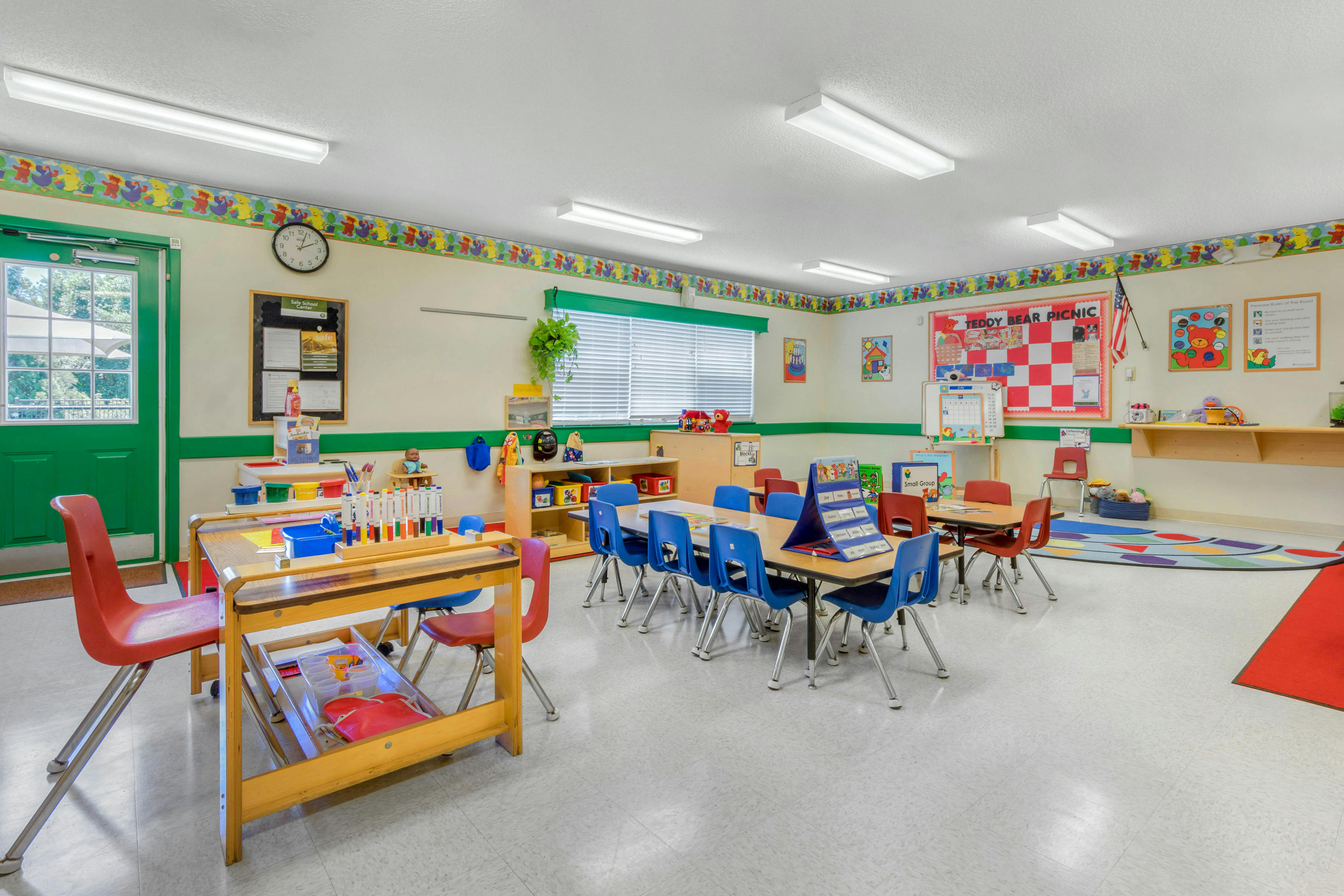 Empty classroom with small tables and chairs, a green door, and bulletin boards