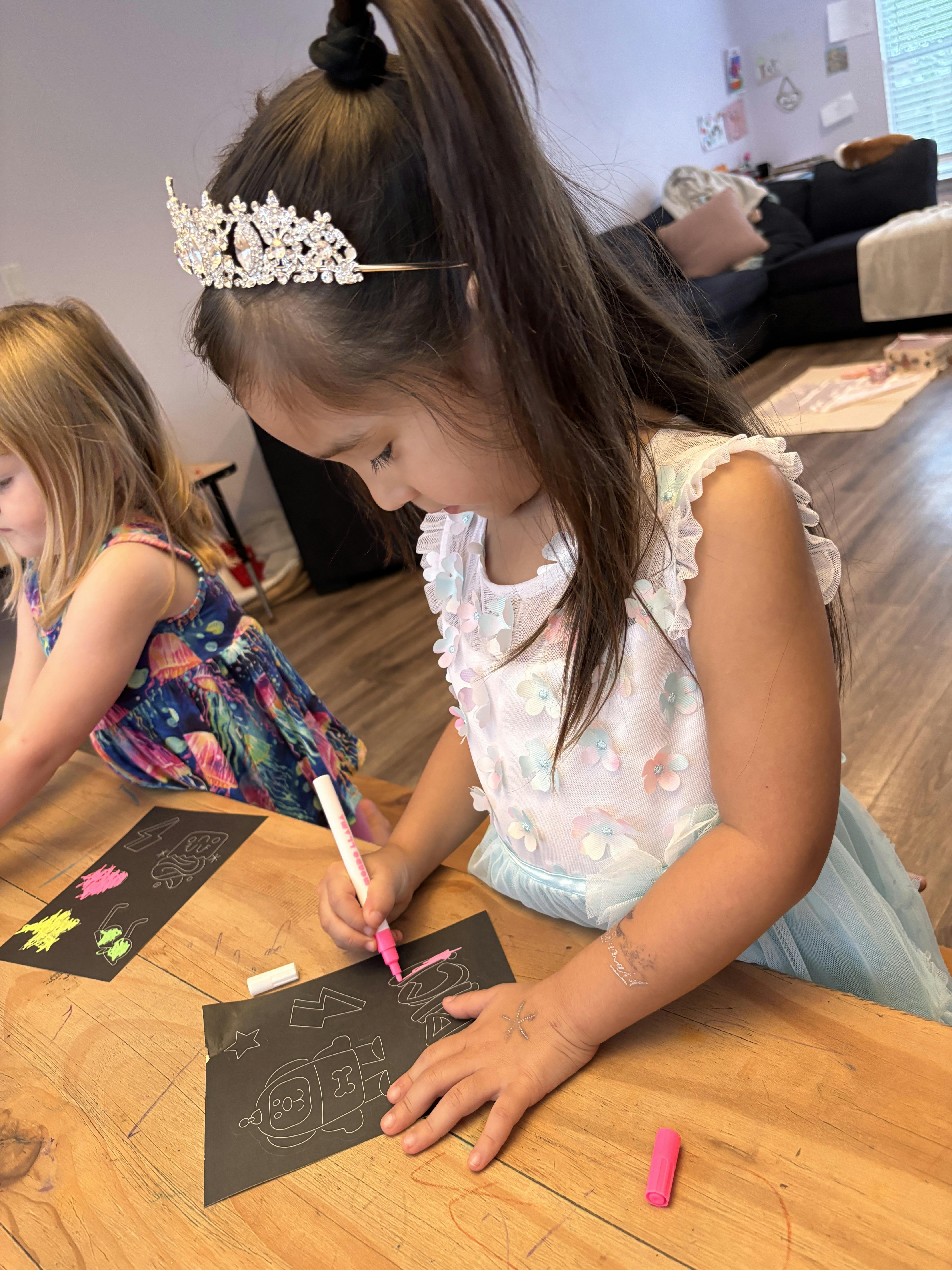 Child wearing a crown drawing on black paper with a pink marker