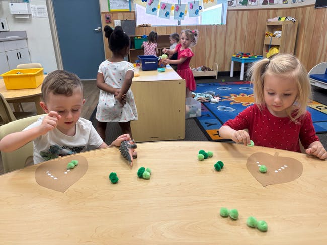 Children playing with green pom-poms at a table in a classroom