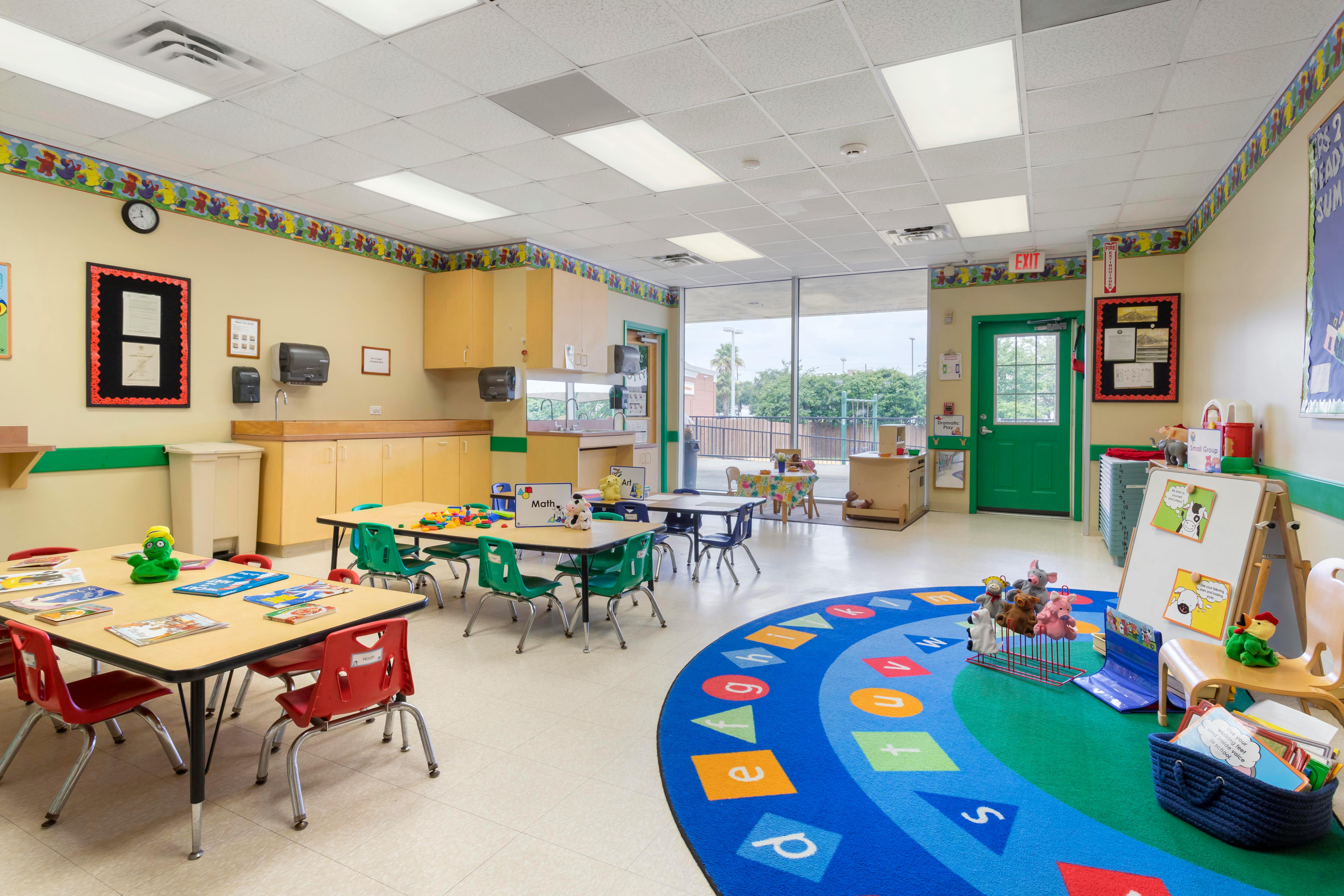 A classroom with small tables and chairs, a play kitchen, and an alphabet rug