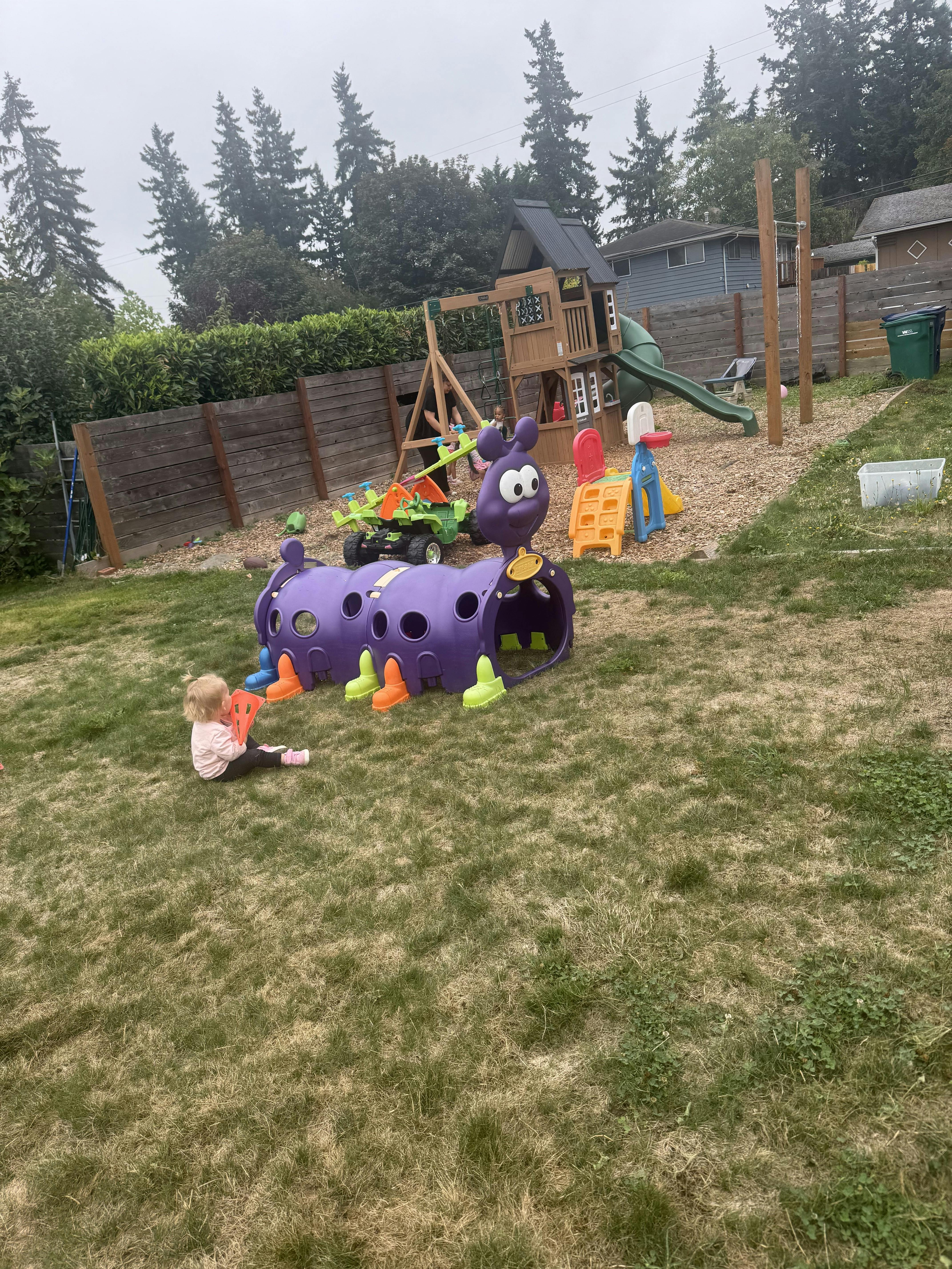Child sitting on grass next to a purple caterpillar play tunnel