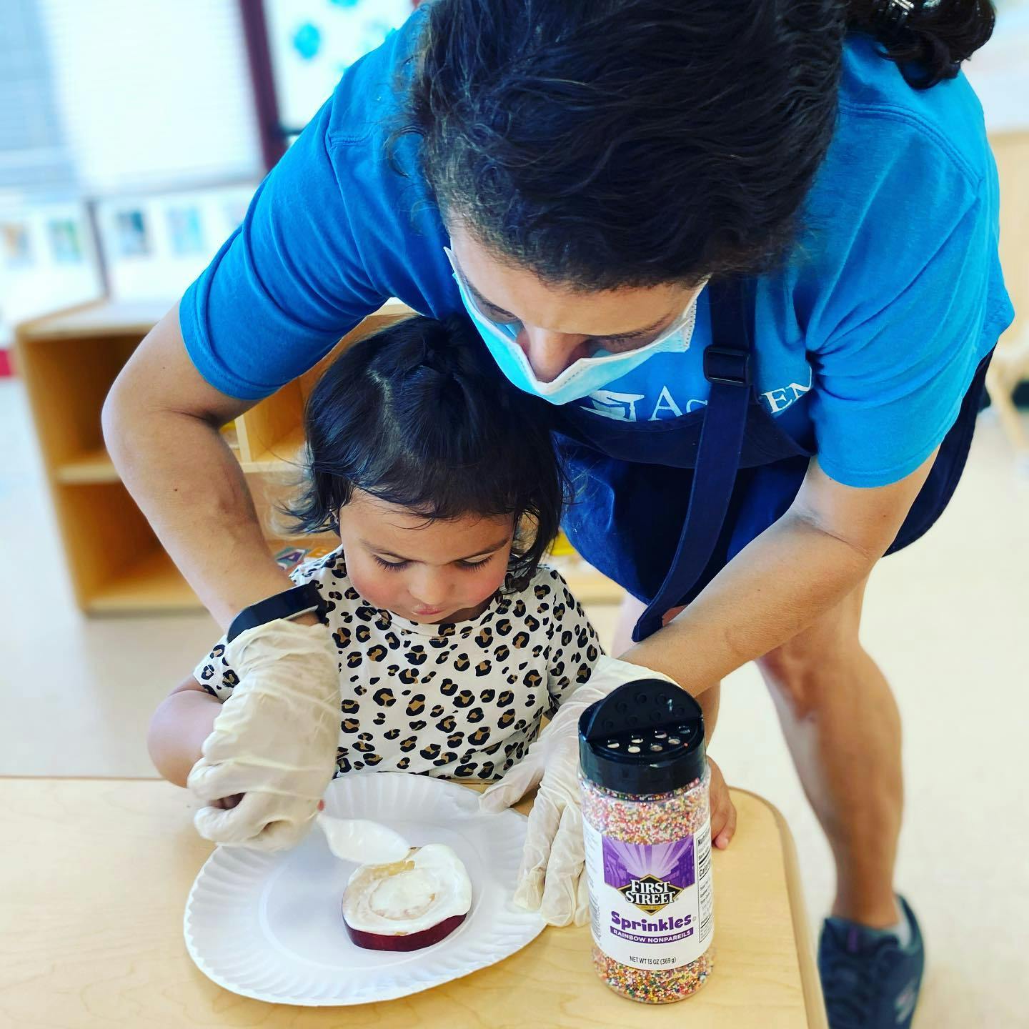 Person in mask helping child with gloves put sprinkles on food