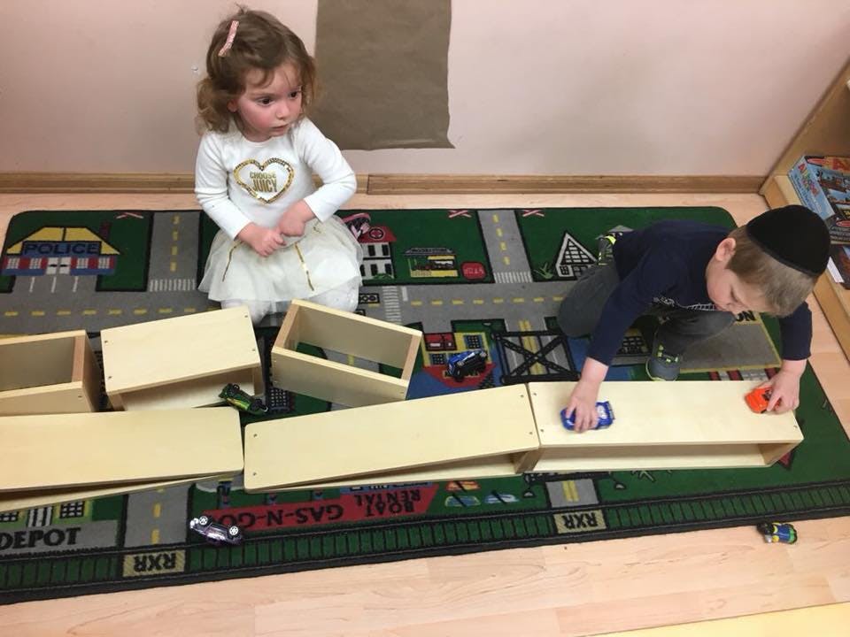 Two children playing with wooden blocks and toy cars on a rug