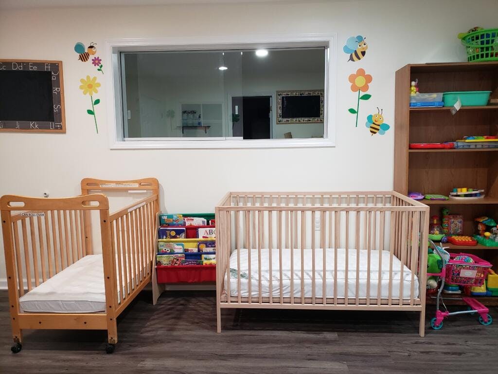 Two empty cribs in a room with a window and shelves