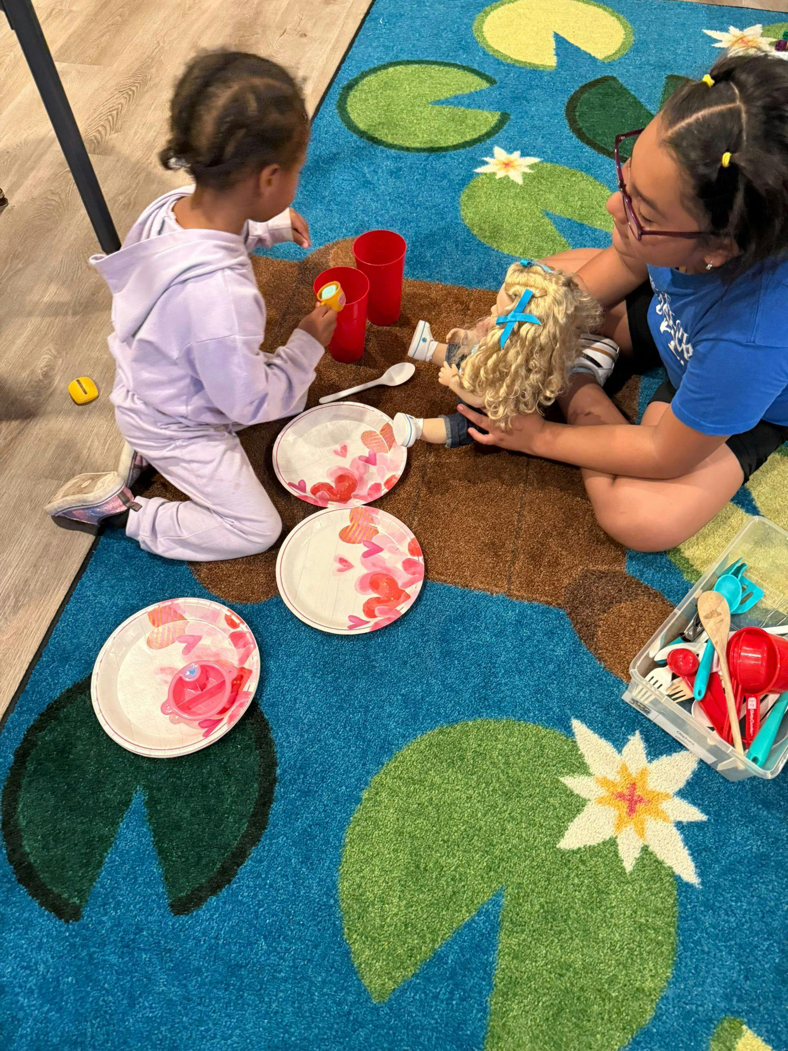 Person and child playing with a doll and toy dishes on a rug