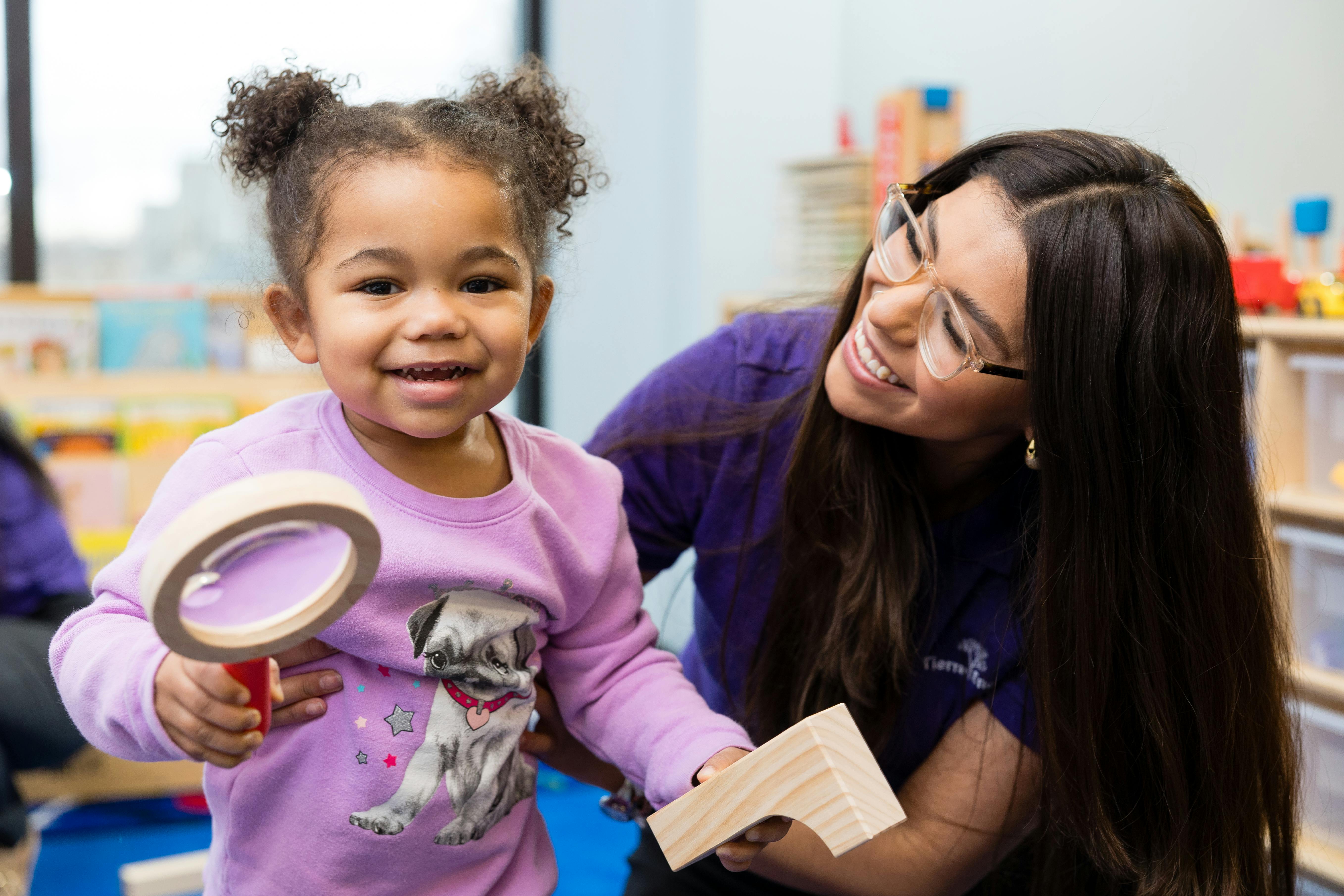 Child holding a magnifying glass next to an adult in a classroom