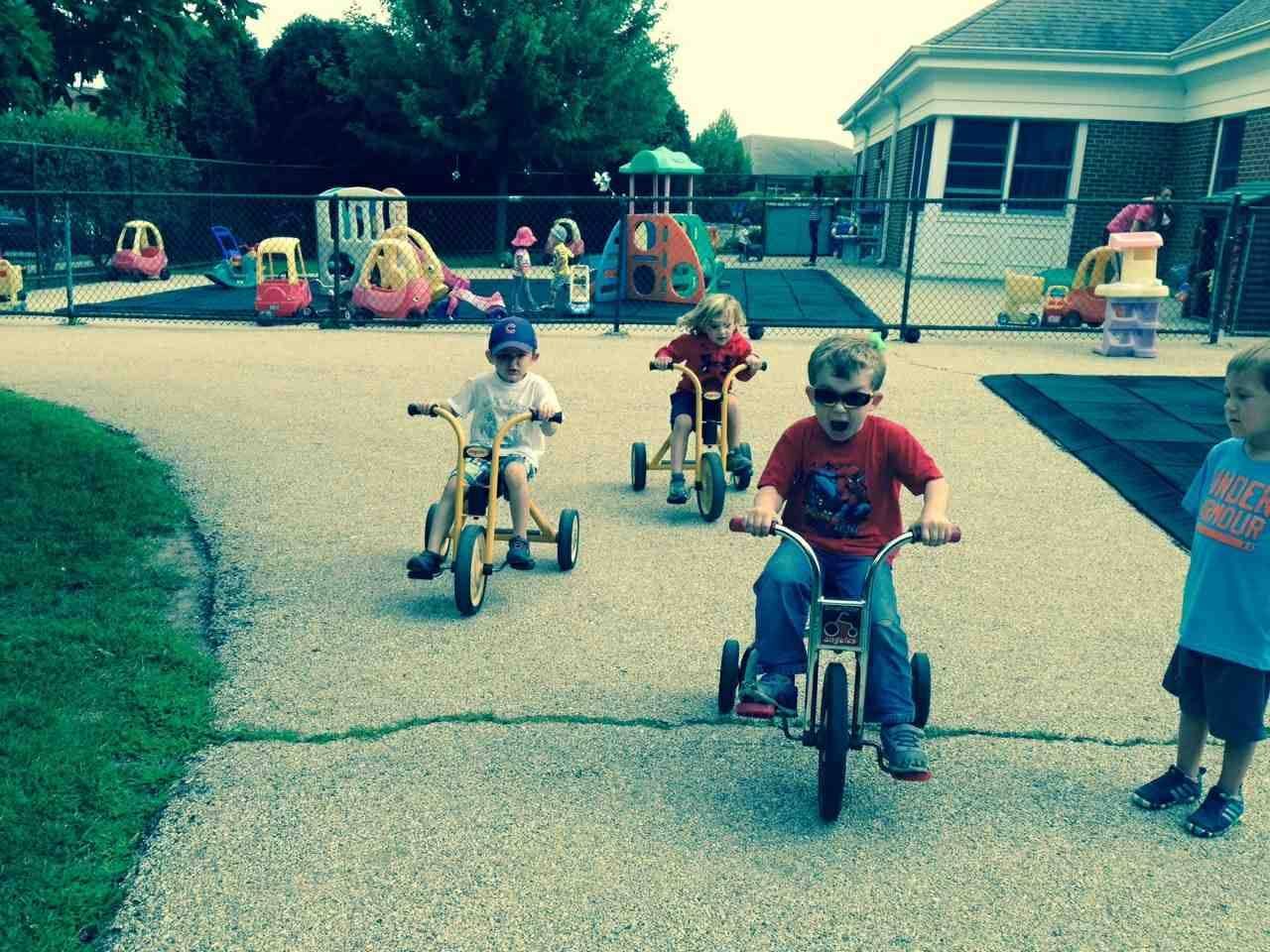 Children riding tricycles on a paved area near a playground