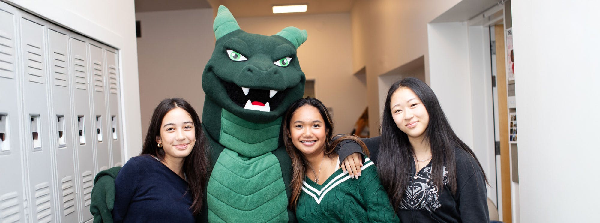 Three teens posing with a green dragon mascot in a school hallway