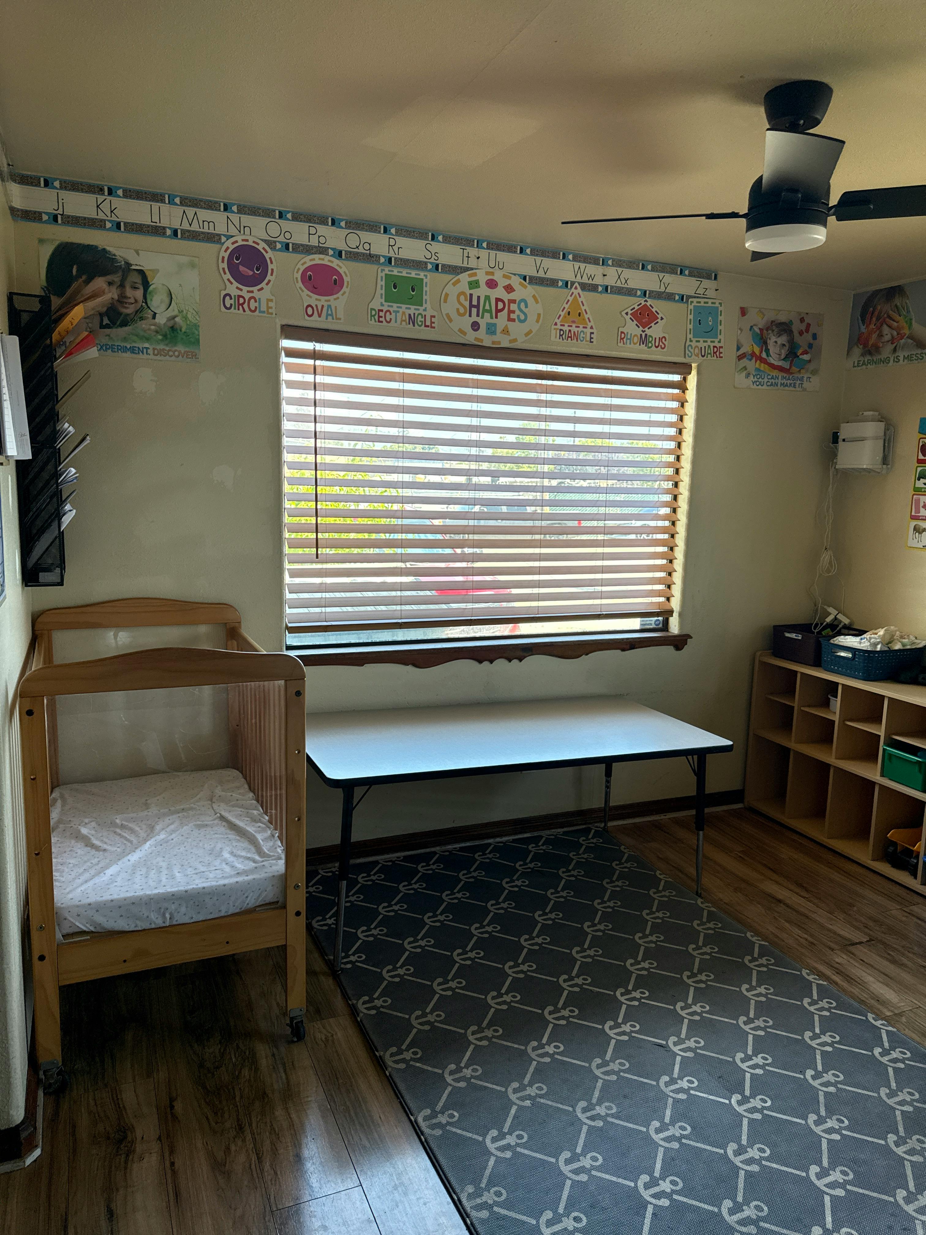Room with classroom decorations, crib, table, ceiling fan, and window blinds