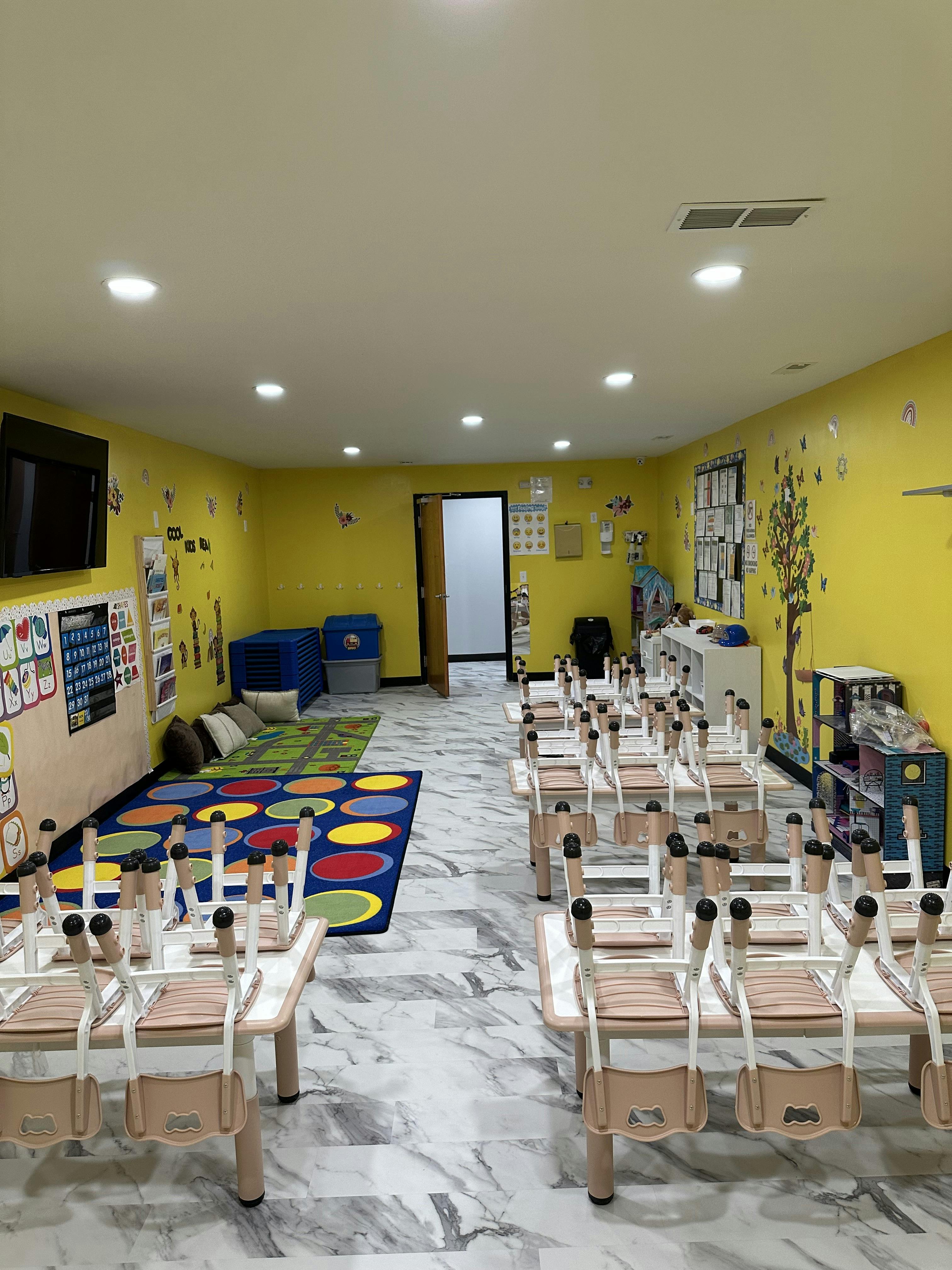 Empty classroom with small tables and chairs, colorful rugs, and yellow walls