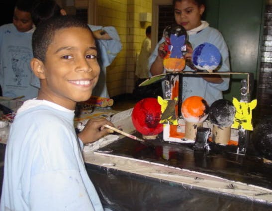 Child painting a craft project, another child in background holding crafts