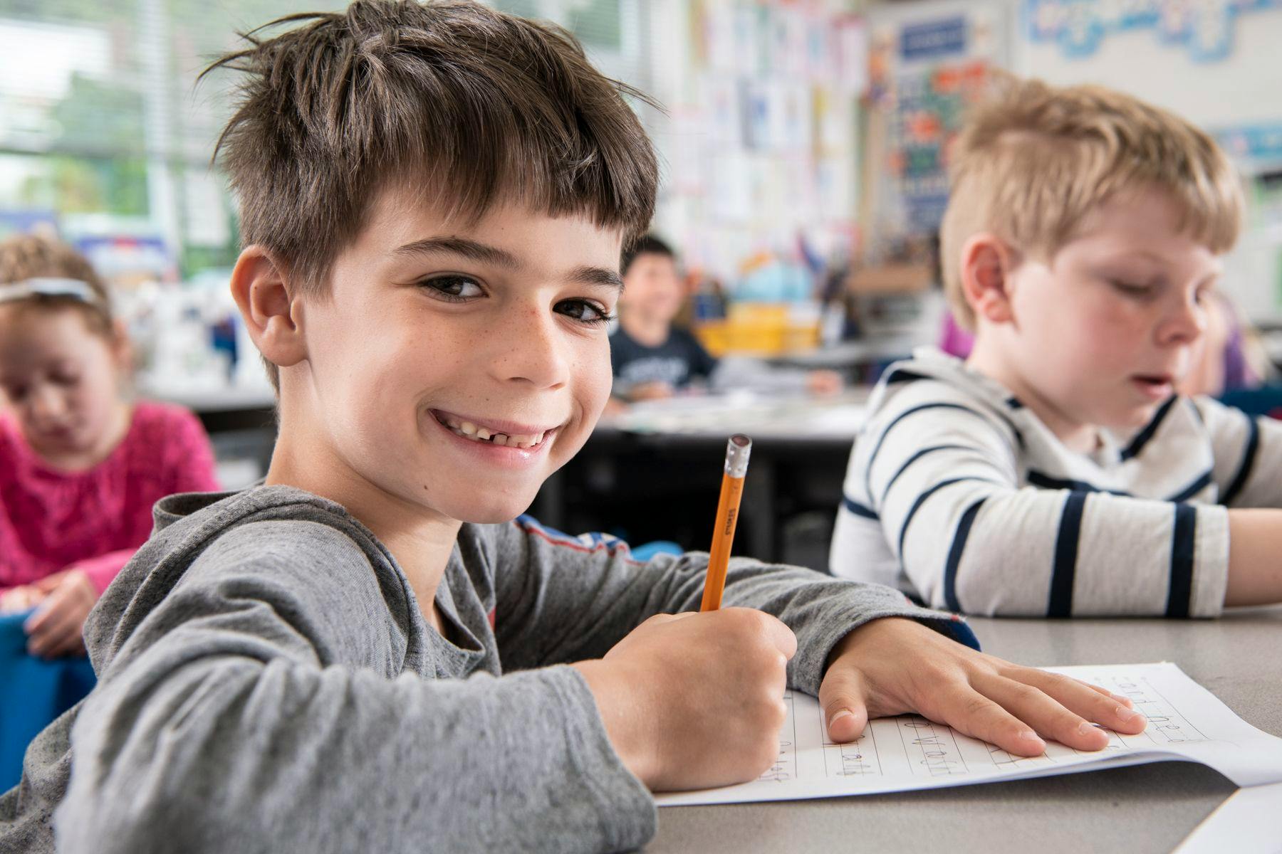 Child writing with a pencil, smiling in a classroom