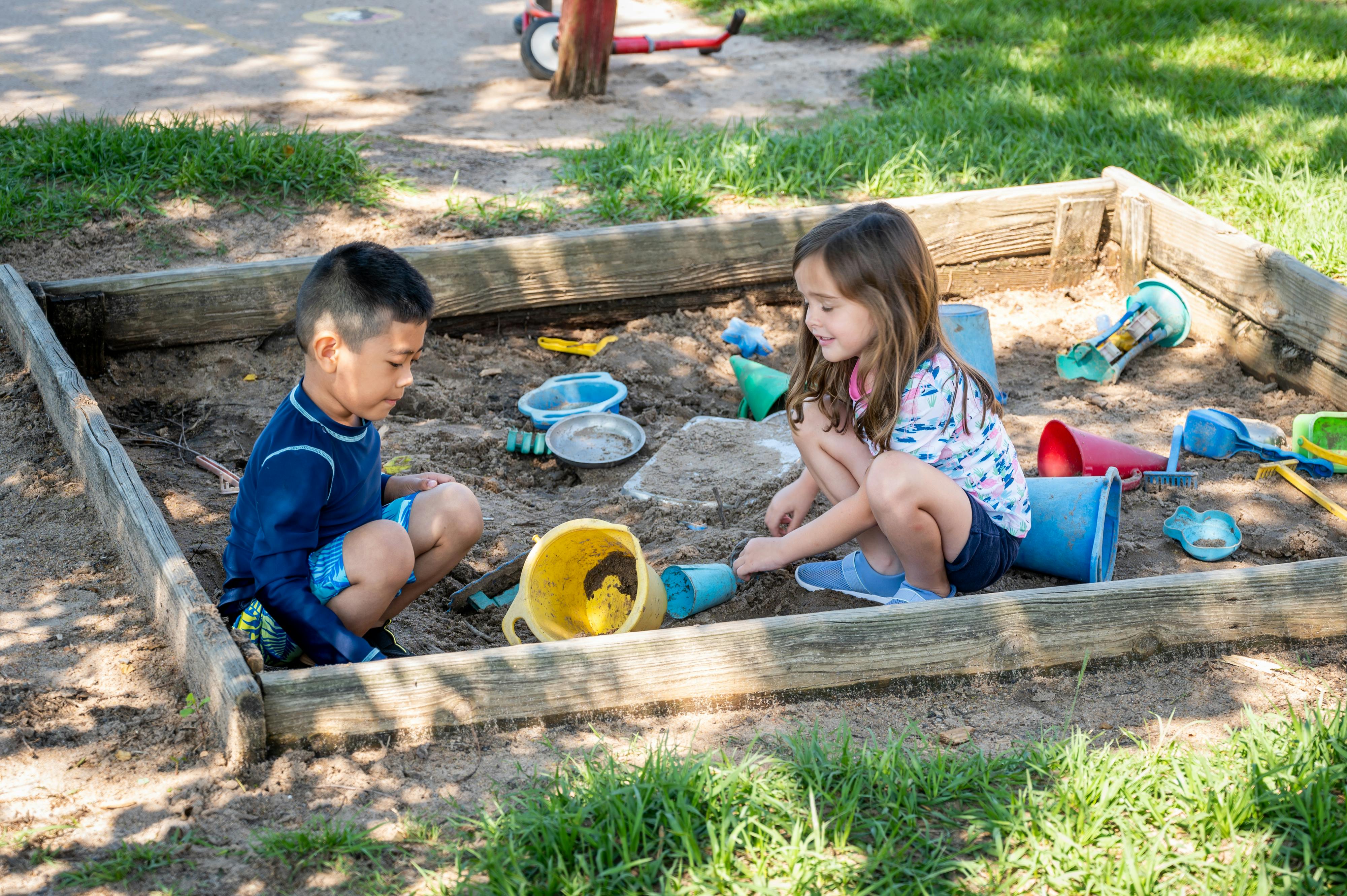 Two children playing with sand and toys in a wooden sandbox